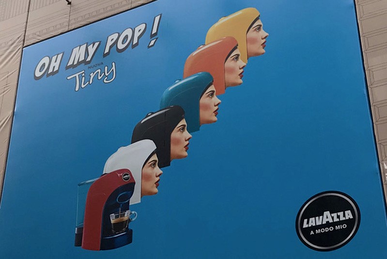 A blue poster showing a 3d computer generated human heads with colorful helmets that look like an espresso machine cover. There is also an espresso machine and the text: Oh my pop! Tiny Lavazza A modo mio..