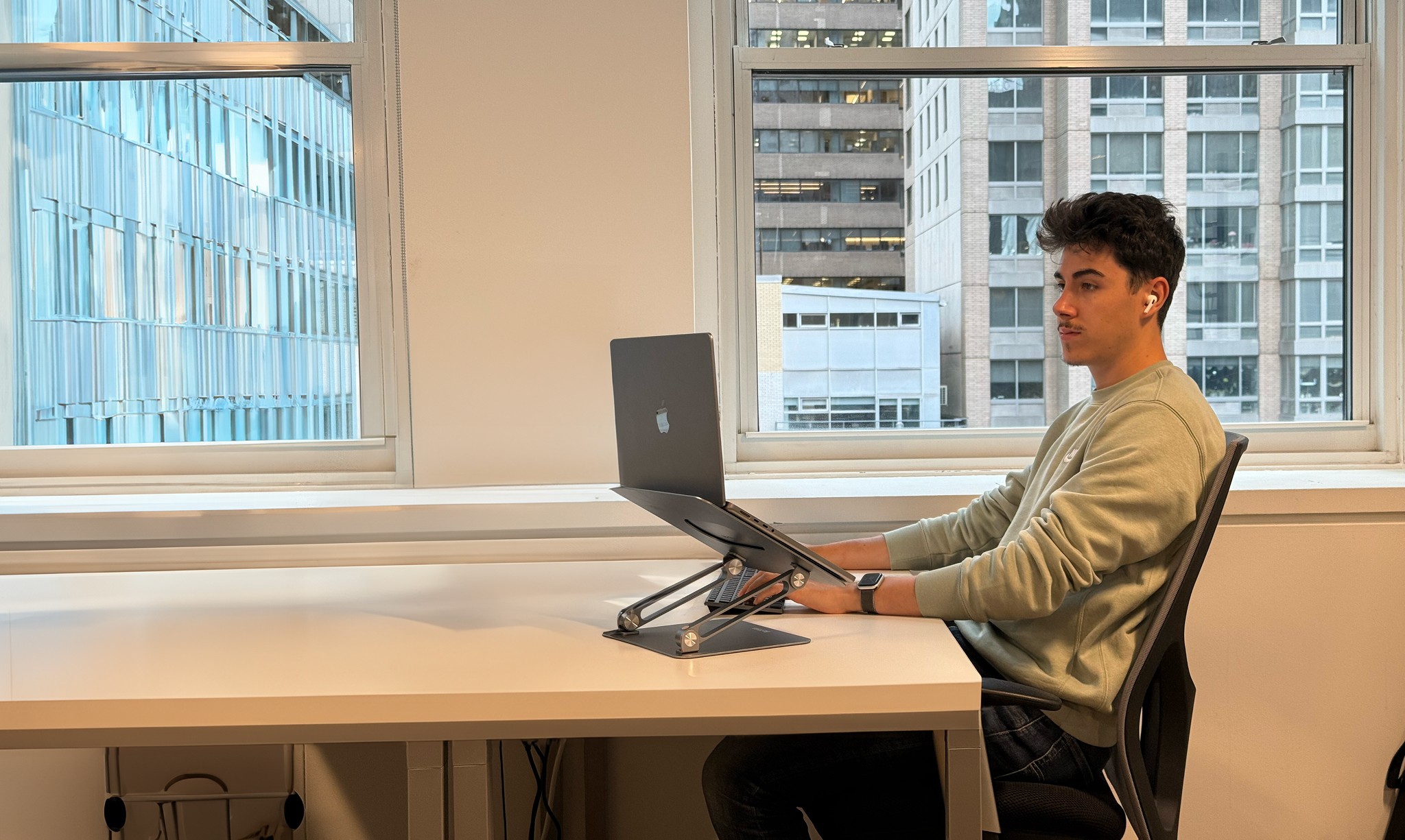 Freelance assis à son bureau dans un open space moderne, travaillant sur un MacBook sur support ergonomique, avec vue sur des immeubles urbains.