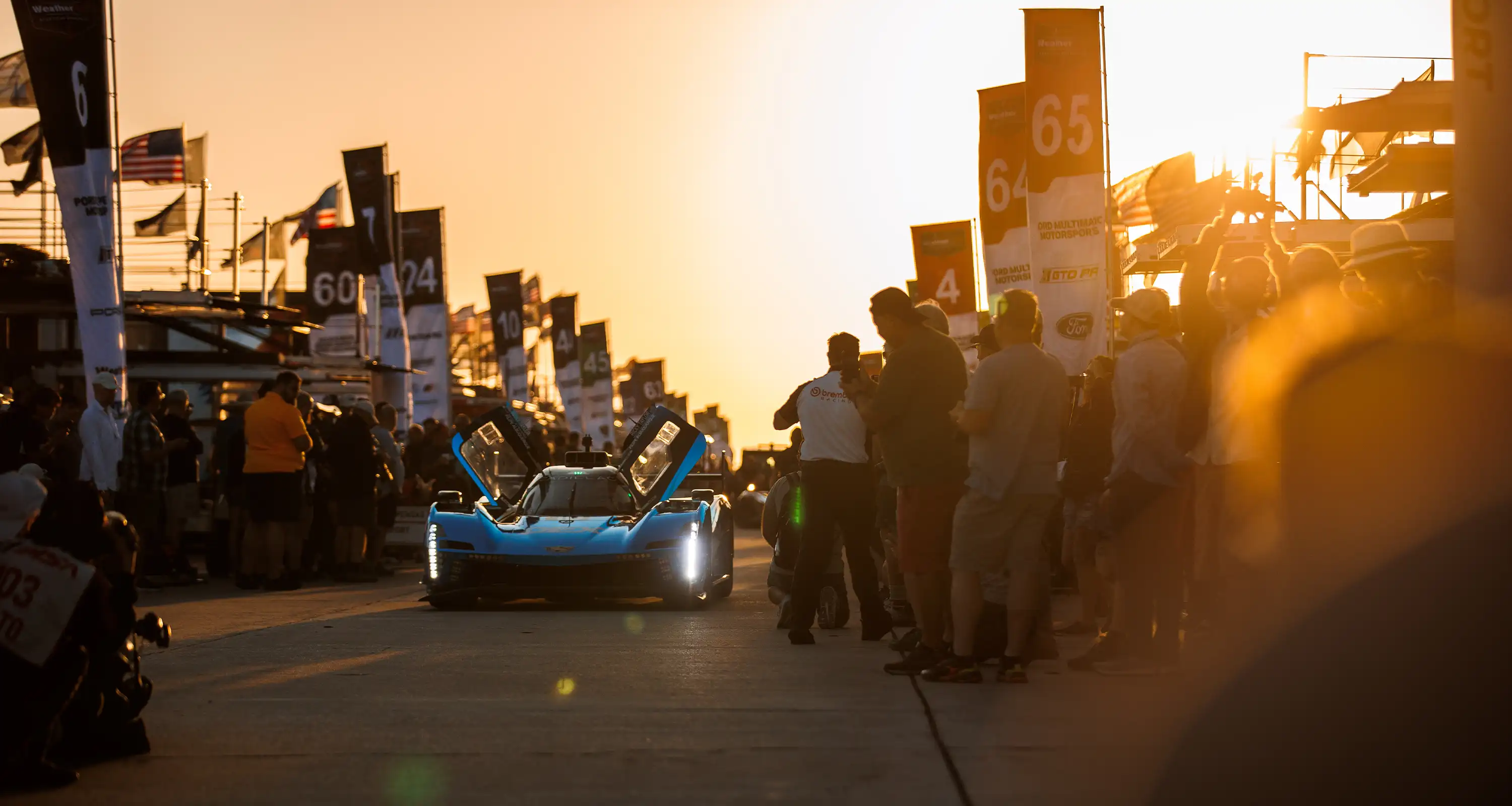 A blue Corvette race car with its doors up drives through the Sebring paddock at sunrise, surrounded by spectators and team members under golden morning light.