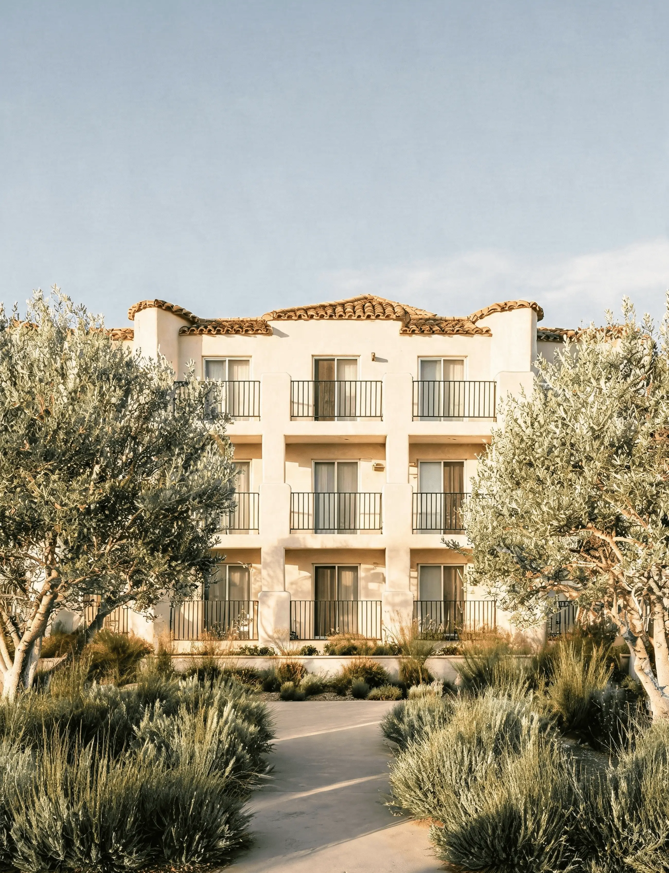 A Mediterranean-style multifamily apartment building with a terracotta tile roof and balconies, surrounded by olive trees.
