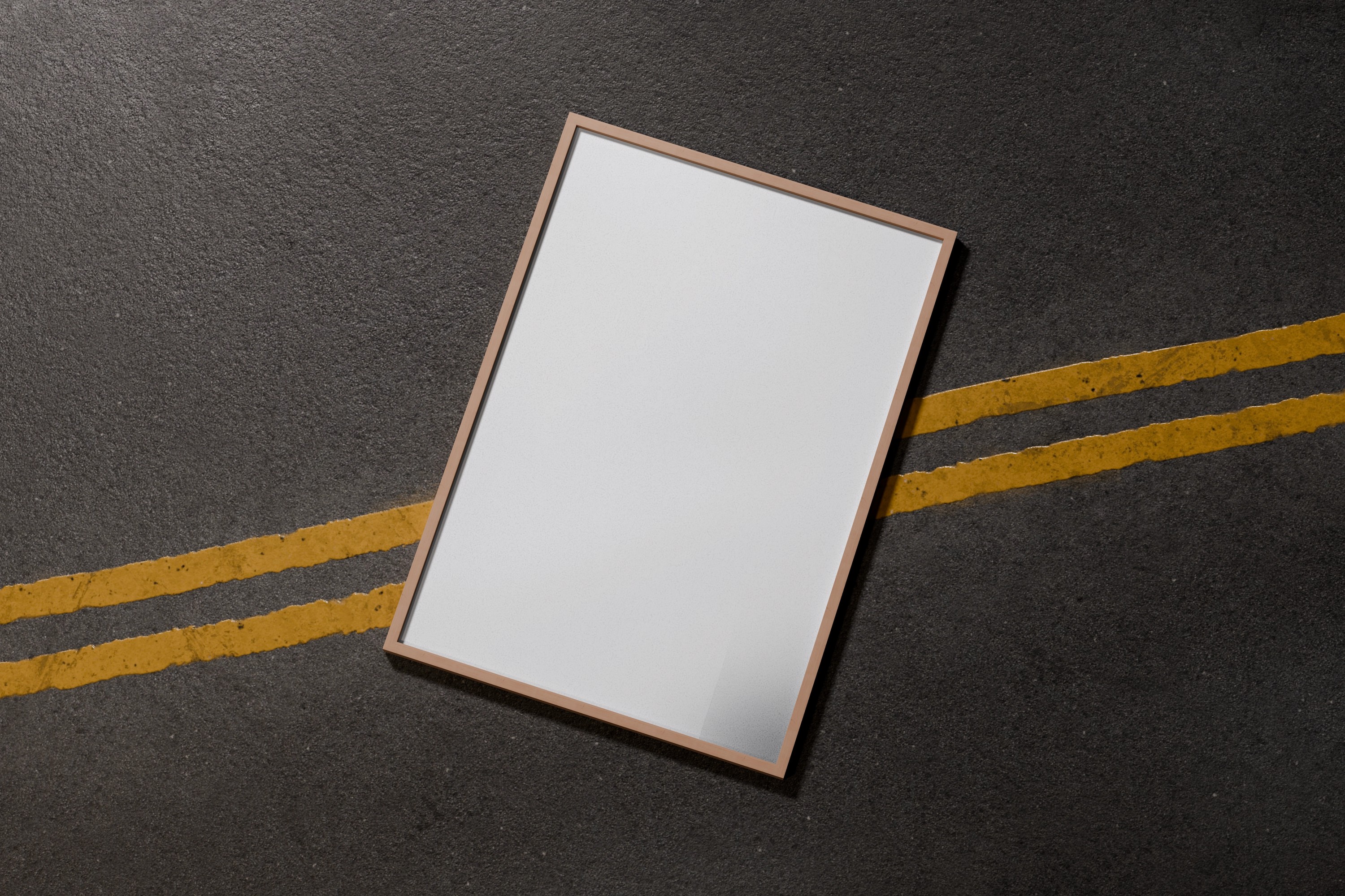 A blank white framed poster lies diagonally on an asphalt surface with two parallel yellow lines, creating a striking urban contrast.