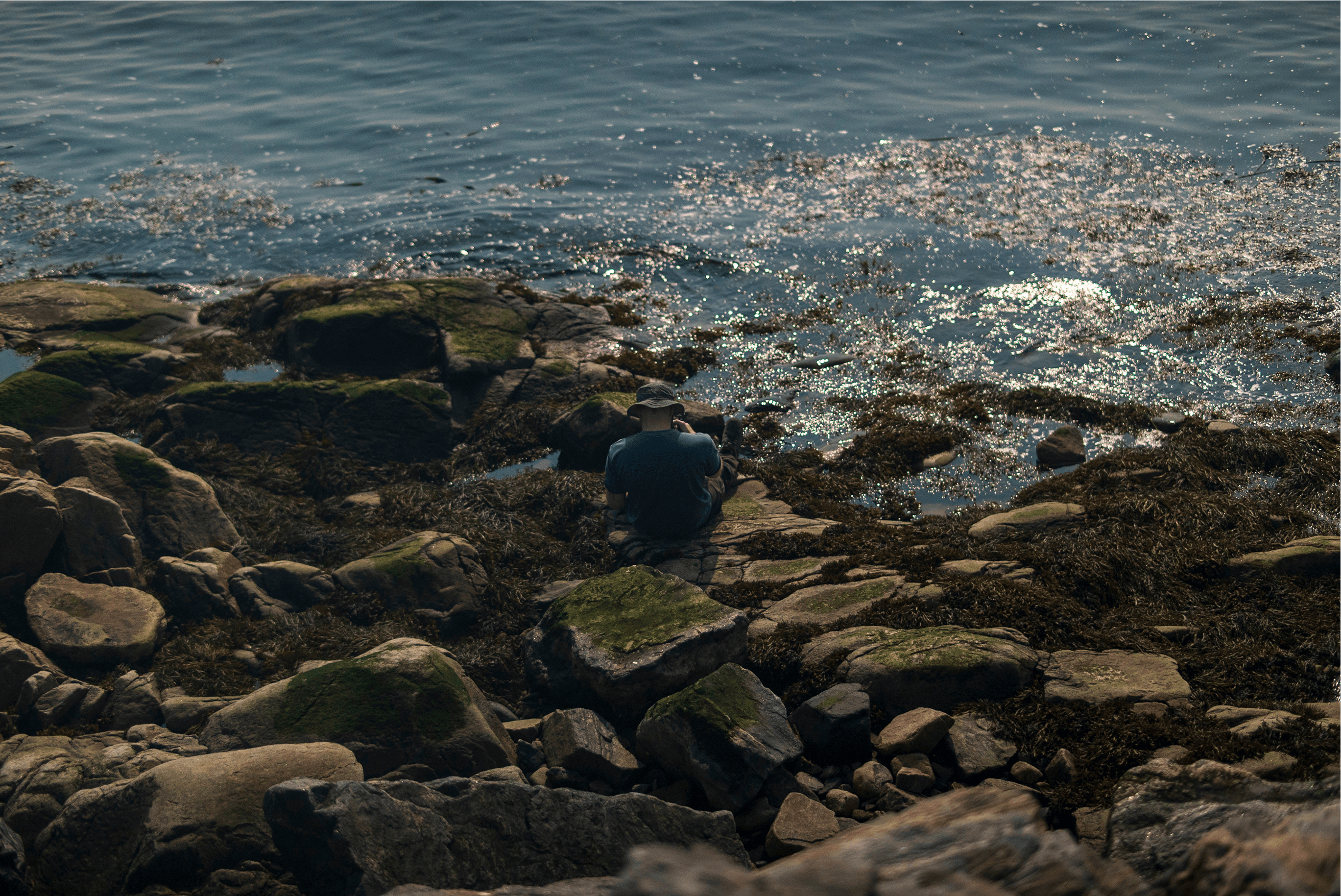 Personne assise sur des rochers couverts d’algues, enregistrant les sons marins face à l’eau scintillante.