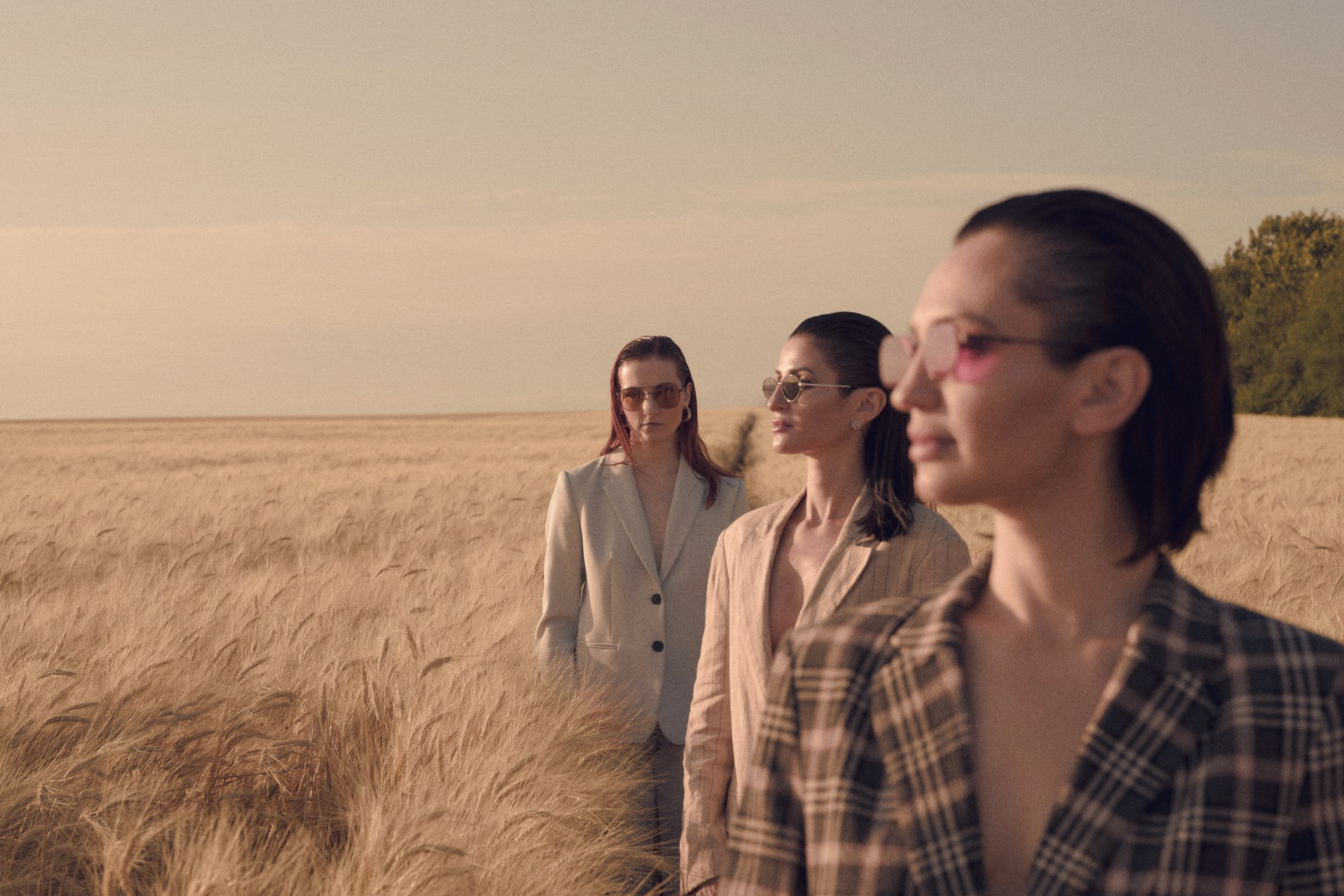 Three women in blazers and sunglasses standing in a vast, golden wheat field under a pale sky at golden hour.
