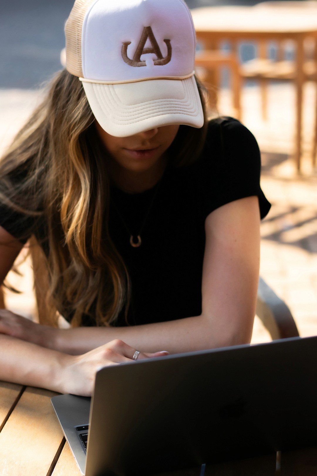 A business owner wearing an anaco creative logo trucker cap, while working on designs on her laptop, sitting outside in the sun at a wooden table.