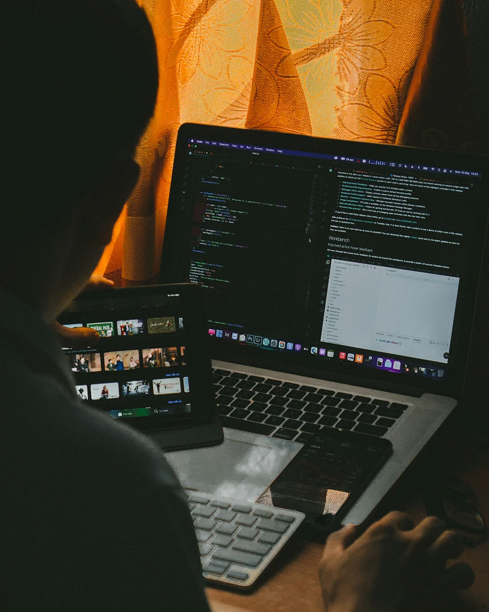 An over-the-shoulder shot of a person working late at night, coding on a laptop while holding a tablet displaying a video streaming app next to a smartphone.