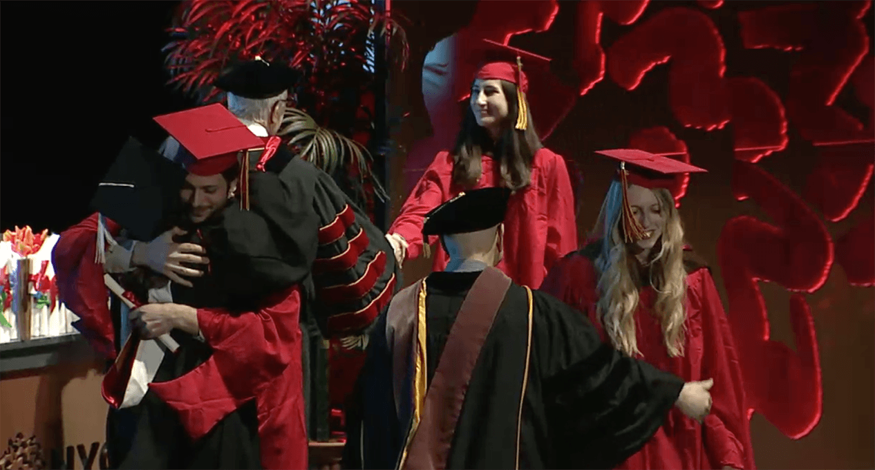 A photo of a couple of grad students and teachers, dressed in SVA color robes, that are hugging and congratulating each other.