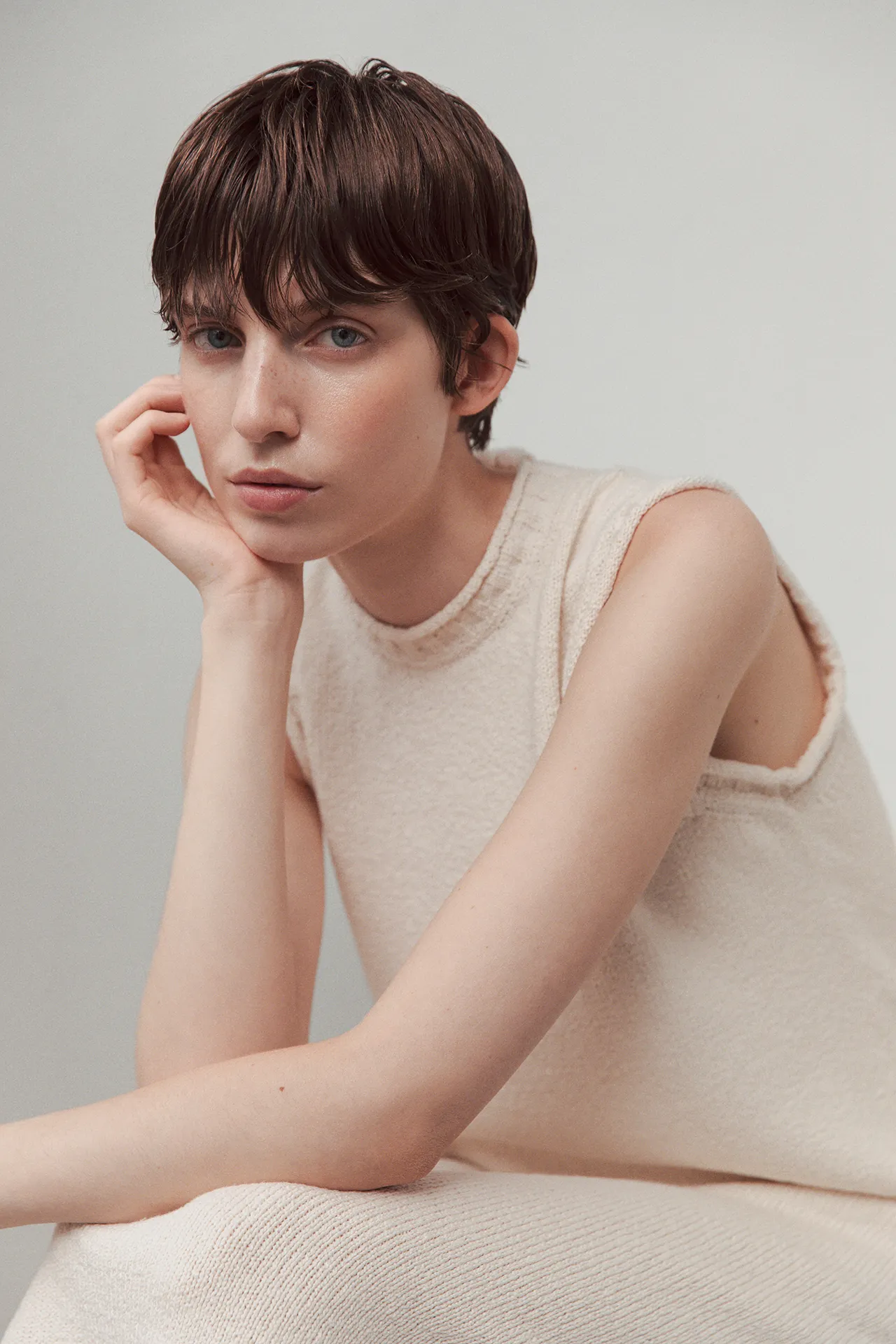 Close-up portrait of a model wearing the Aida Barni SS26 collection cream sleeveless knit dress, resting her face on her hand.