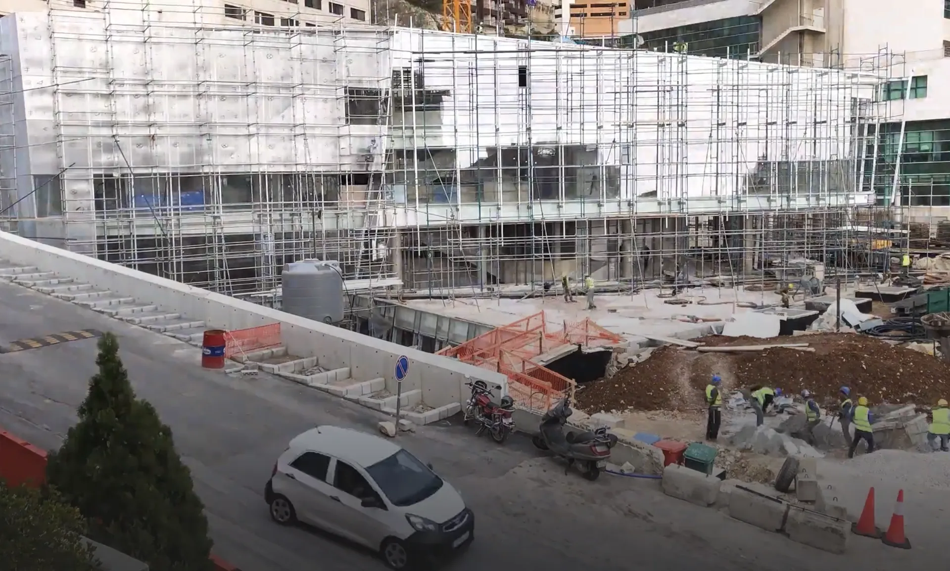 Modern architectural workspace with large floor-to-ceiling windows, surrounded by trees, and desks filled with design materials and books. Two blurred figures are walking through the space.