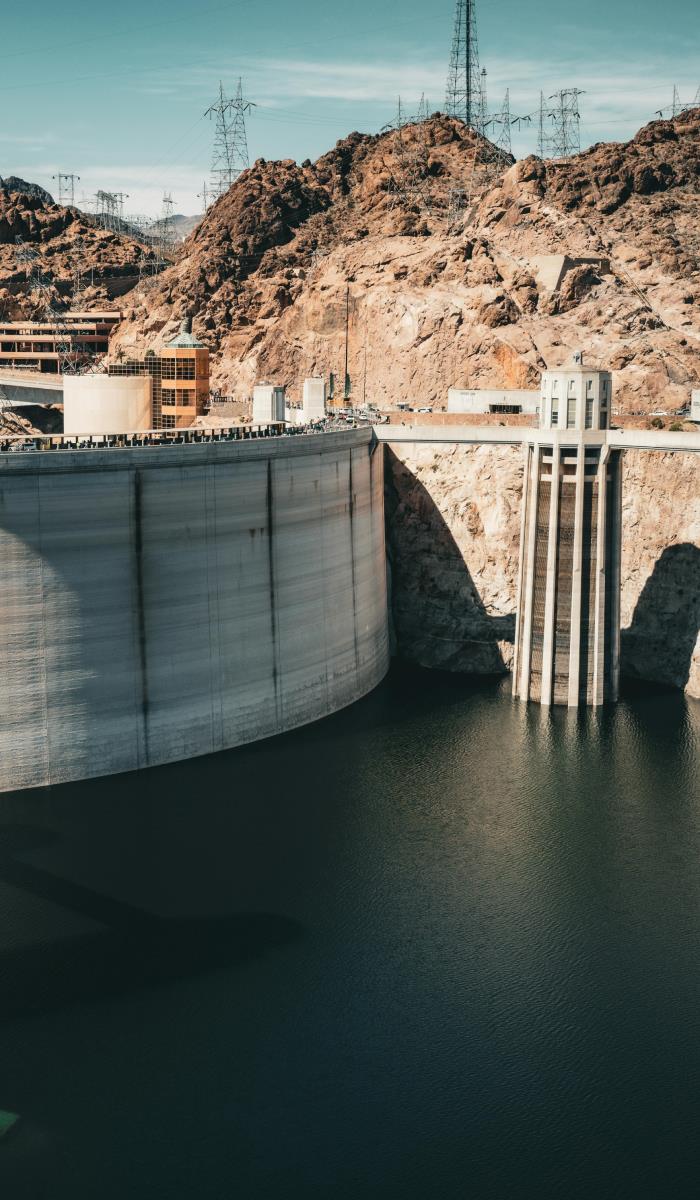 A massive concrete dam stretches across calm water, set against rugged, rocky cliffs under a clear sky. Power lines are visible above, emphasizing its grandeur.