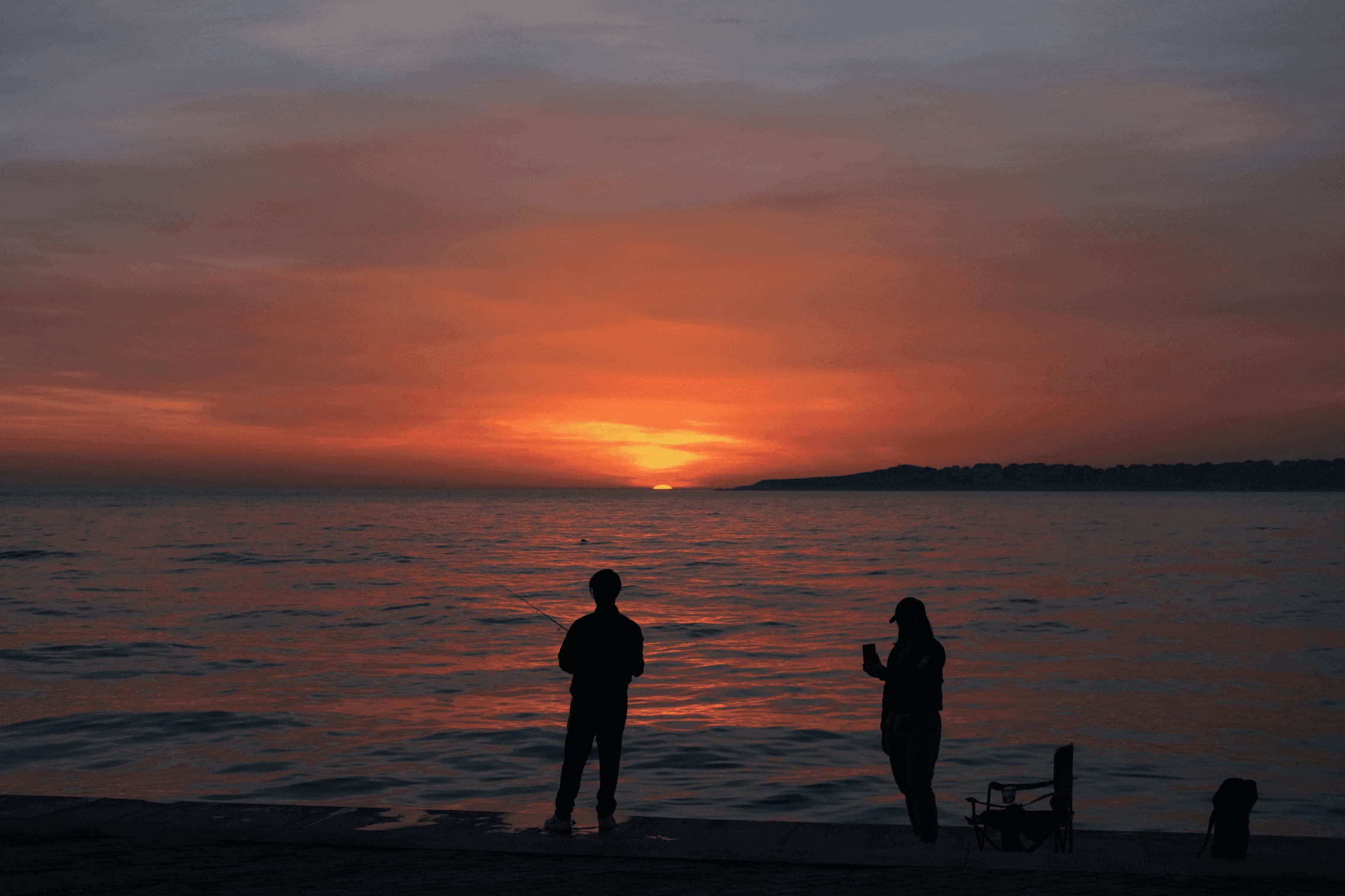 Two silhouettes stand on the shore, watching a vibrant sunset over the water, with soft clouds in the sky.
