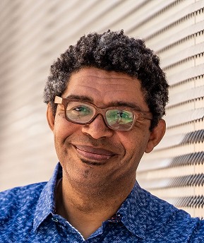 A person with curly hair and glasses smiles while standing in front of a metal striped wall, wearing a blue patterned shirt.