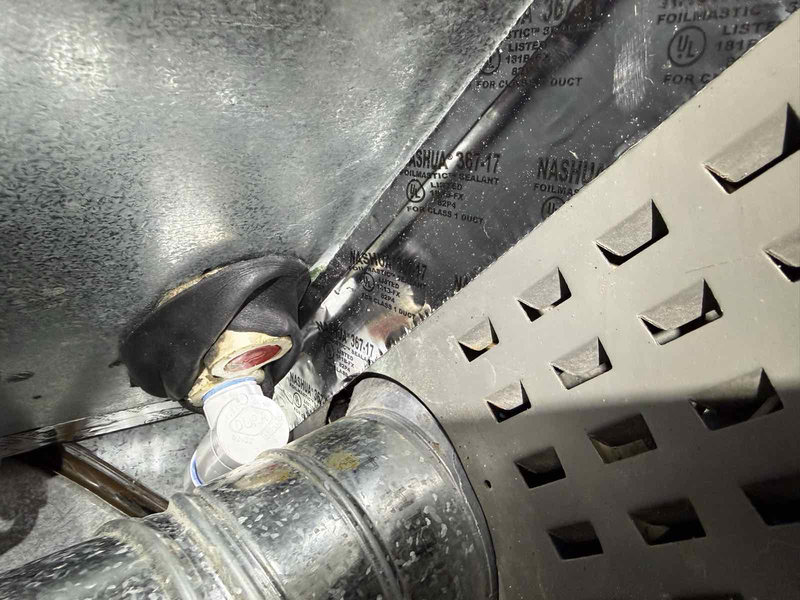 A close view of a corroded brass compression fitting on a condensate drain, packed with black mastic where it passes through sheet metal.