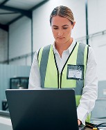 A person in a high-visibility safety vest is working on a laptop inside a modern industrial facility, with equipment and machinery visible in the background.