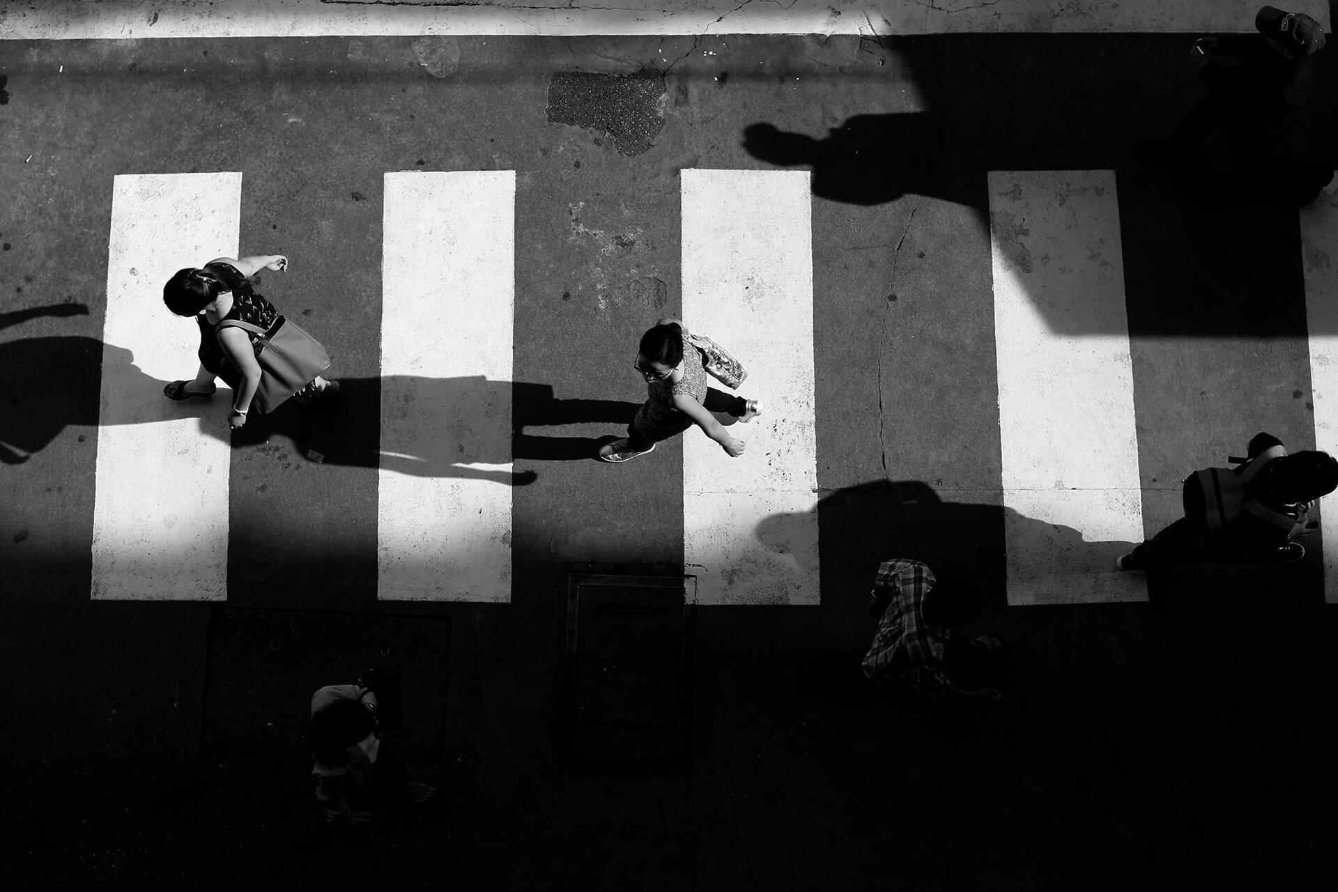 A black-and-white overhead photo captures people walking across a striped crosswalk, casting long, sharp shadows on the pavement. The mix of light and dark areas highlights the movement and contrast in an urban setting.