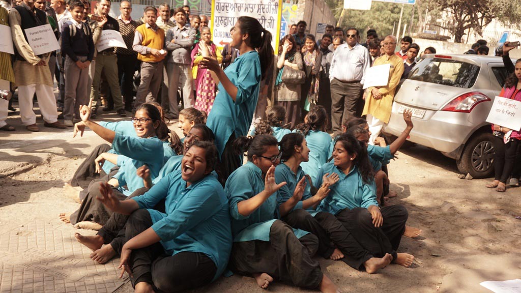 A smiling young man with crossed arms, wearing a plaid shirt and white t-shirt, poses against a dark background.