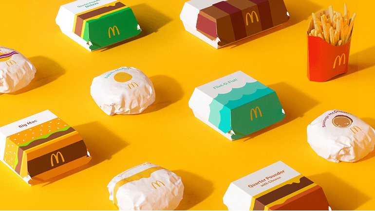 McDonald’s food packaging arranged on a bright yellow background, including wrapped sandwiches and branded boxes with the McDonald’s logo.