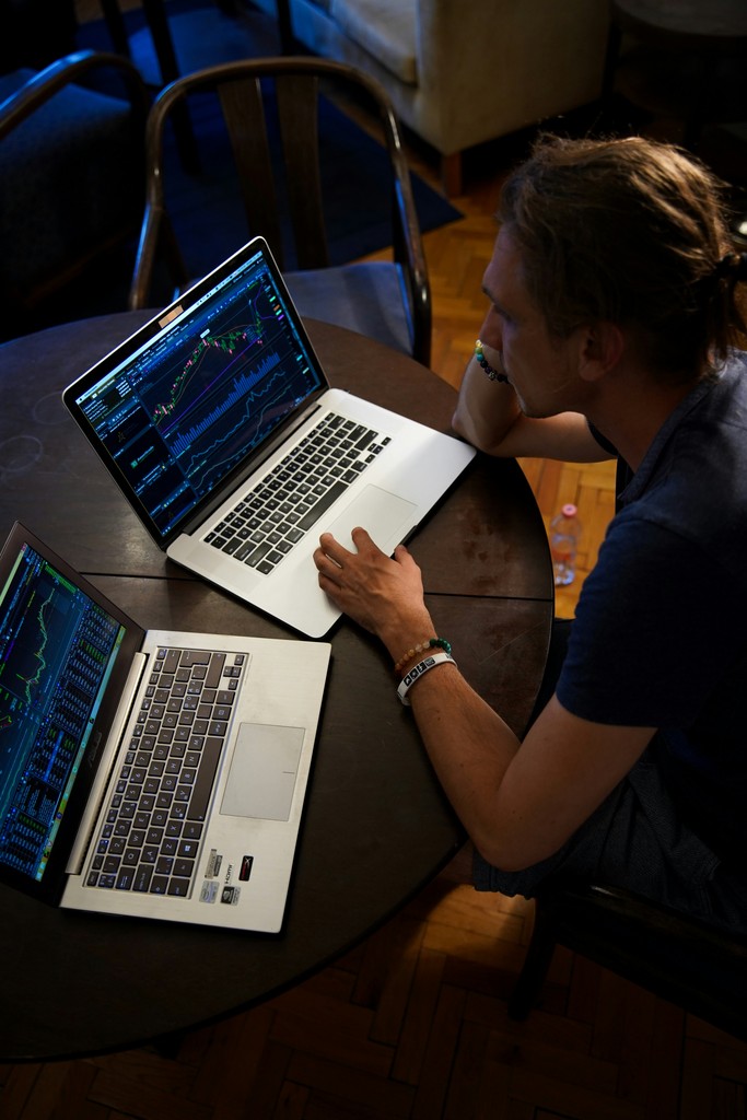 A person sits at a round table using two laptops displaying complex financial charts and graphs. The scene, set in a dimly lit room, emphasizes focus and analysis, suggesting activities related to stock trading, financial analysis, or data monitoring.