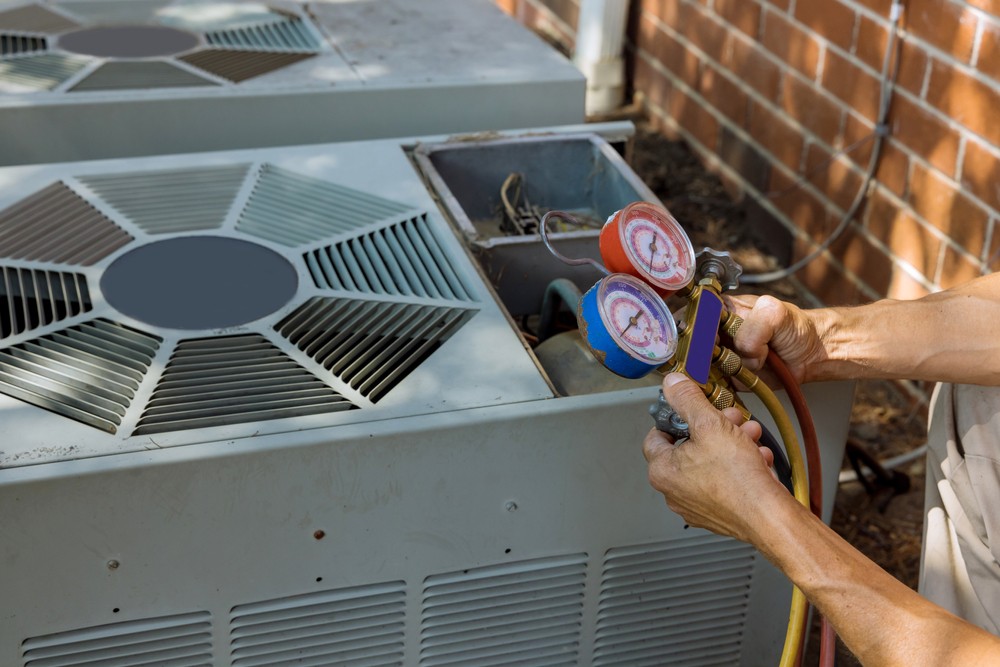 A technician's hands holding a pair of HVAC manifold gauges connected to an outdoor air conditioning unit. The unit’s fan grilles are visible against a brick wall backdrop. These tools are essential for diagnosing and servicing the system, ensuring proper air conditioning installation in Asheville.