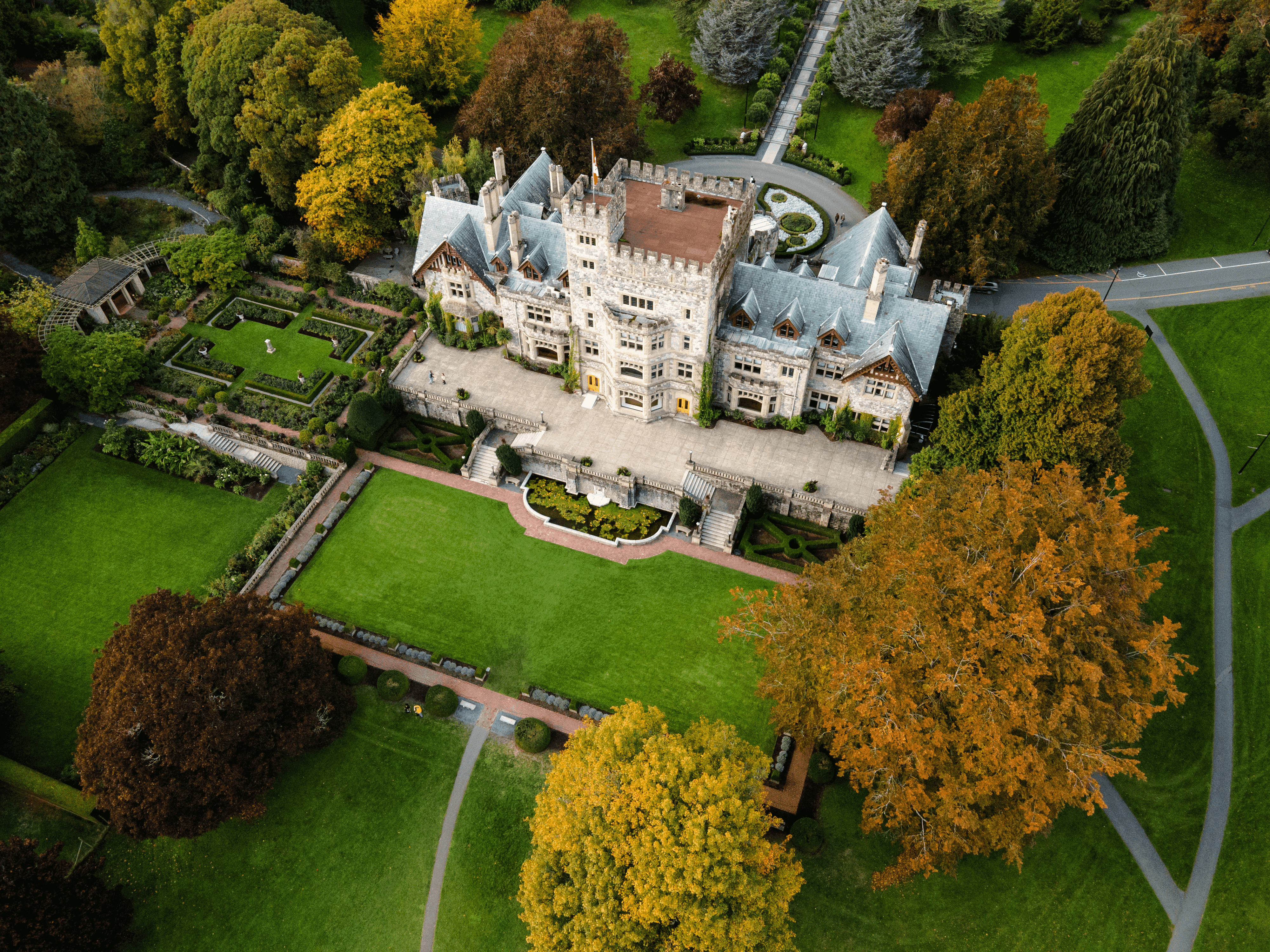 Aerial view of a large historic castle-style mansion surrounded by manicured gardens, green lawns, winding paths, and autumn-colored trees.