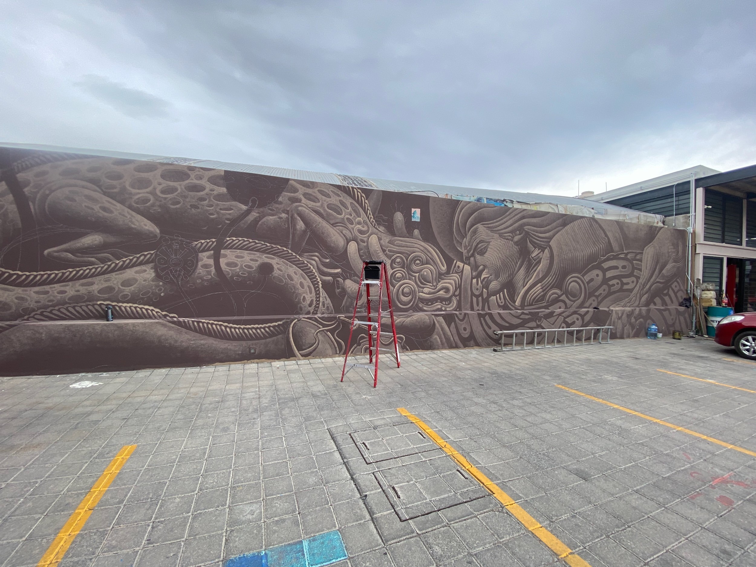 The finished mural stretches the length of a grey wall above an empty paved lot, a red stepladder in front.