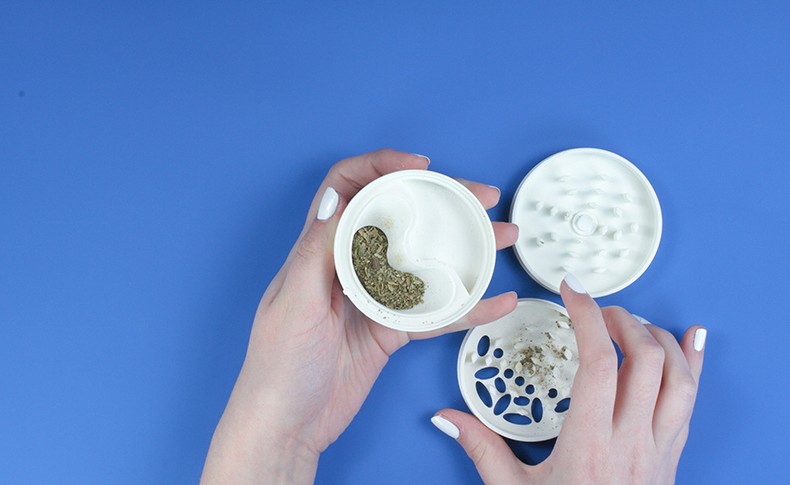 Two hands with white nail polish are holding open two white plastic containers against a solid blue background. One container holds a green, granular substance. The other container has a pattern of blue ovals on its base and contains some loose material. The containers are positioned diagonally.