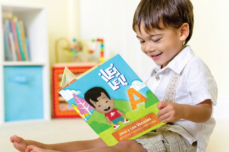 A young child with short dark hair, wearing a white short-sleeved shirt and light-colored shorts, is seated barefoot and holding a book. The book has a light blue cover and features illustrations of a boy and a large letter "A." The title "Jaul y las vocales" is visible on the cover, along with the author's name, "Francisco J. Hernandez." The child is looking down at the book with concentration. The background includes a light-colored wall, a white shelving unit with books, and a colorful toy.