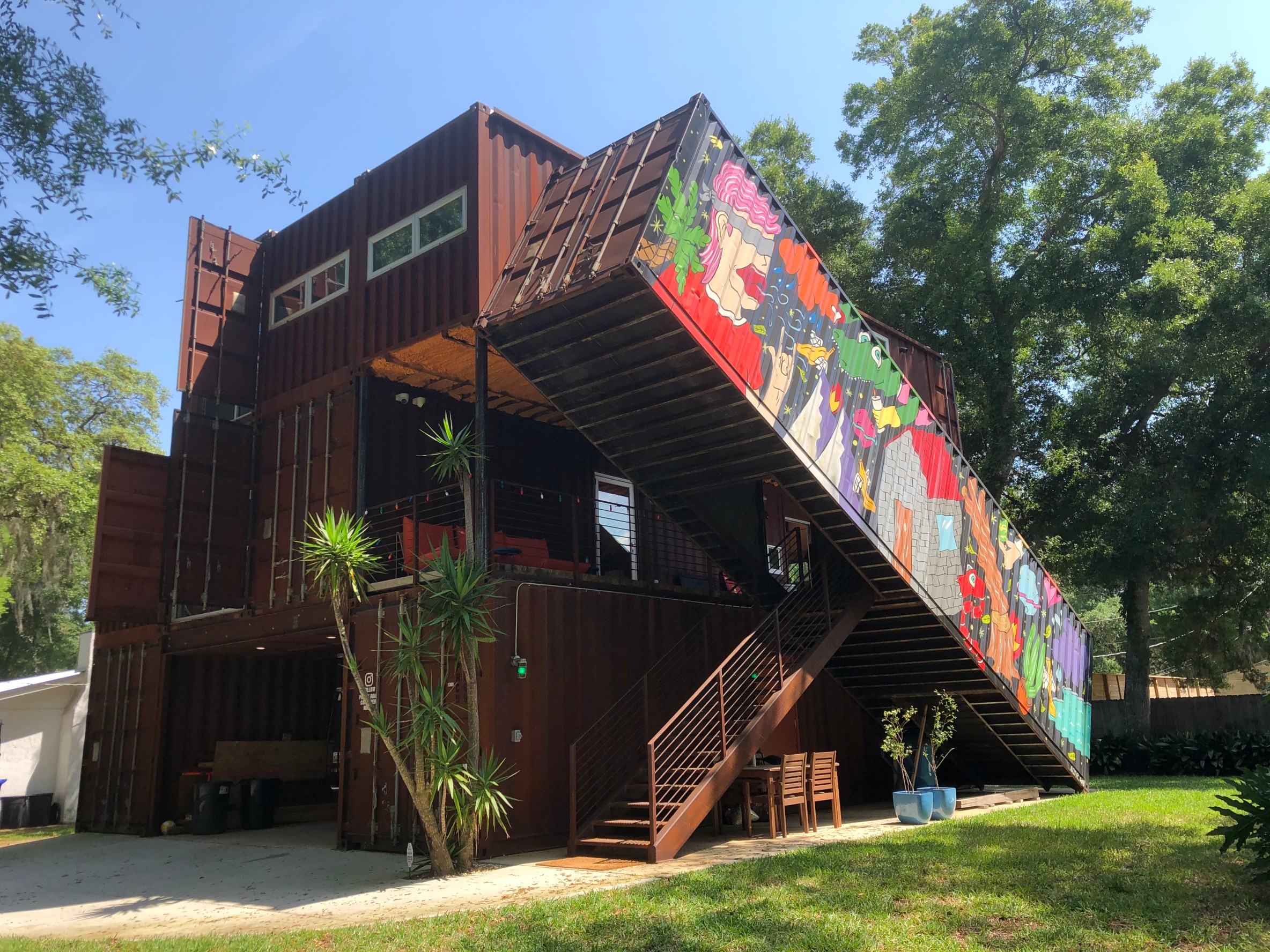 A unique Airbnb rental made from stacked shipping containers, featuring a colorful mural along the diagonal staircase and surrounded by lush trees and plants.