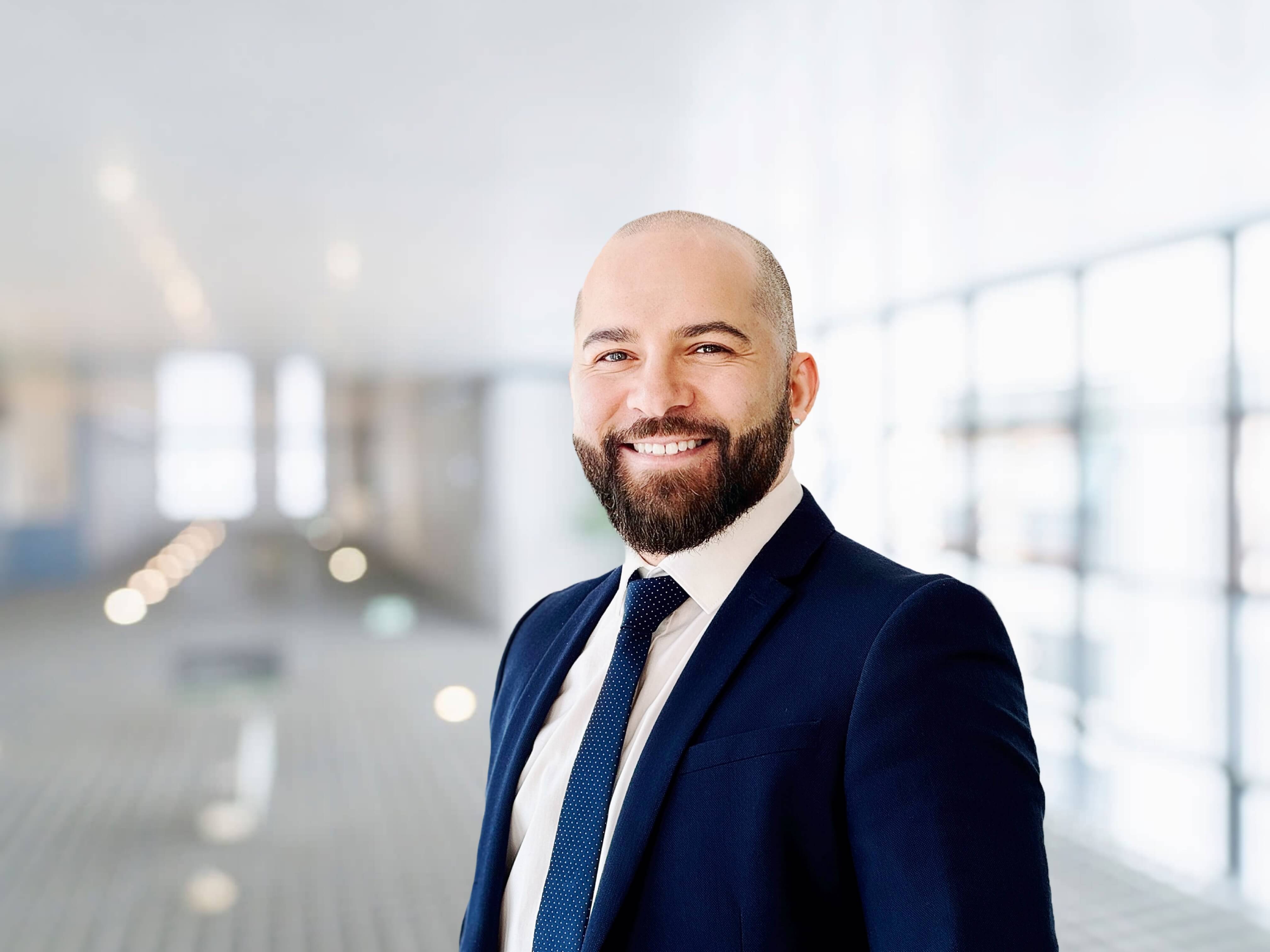 A smiling man with a beard in a suit stands in a bright, modern office space.