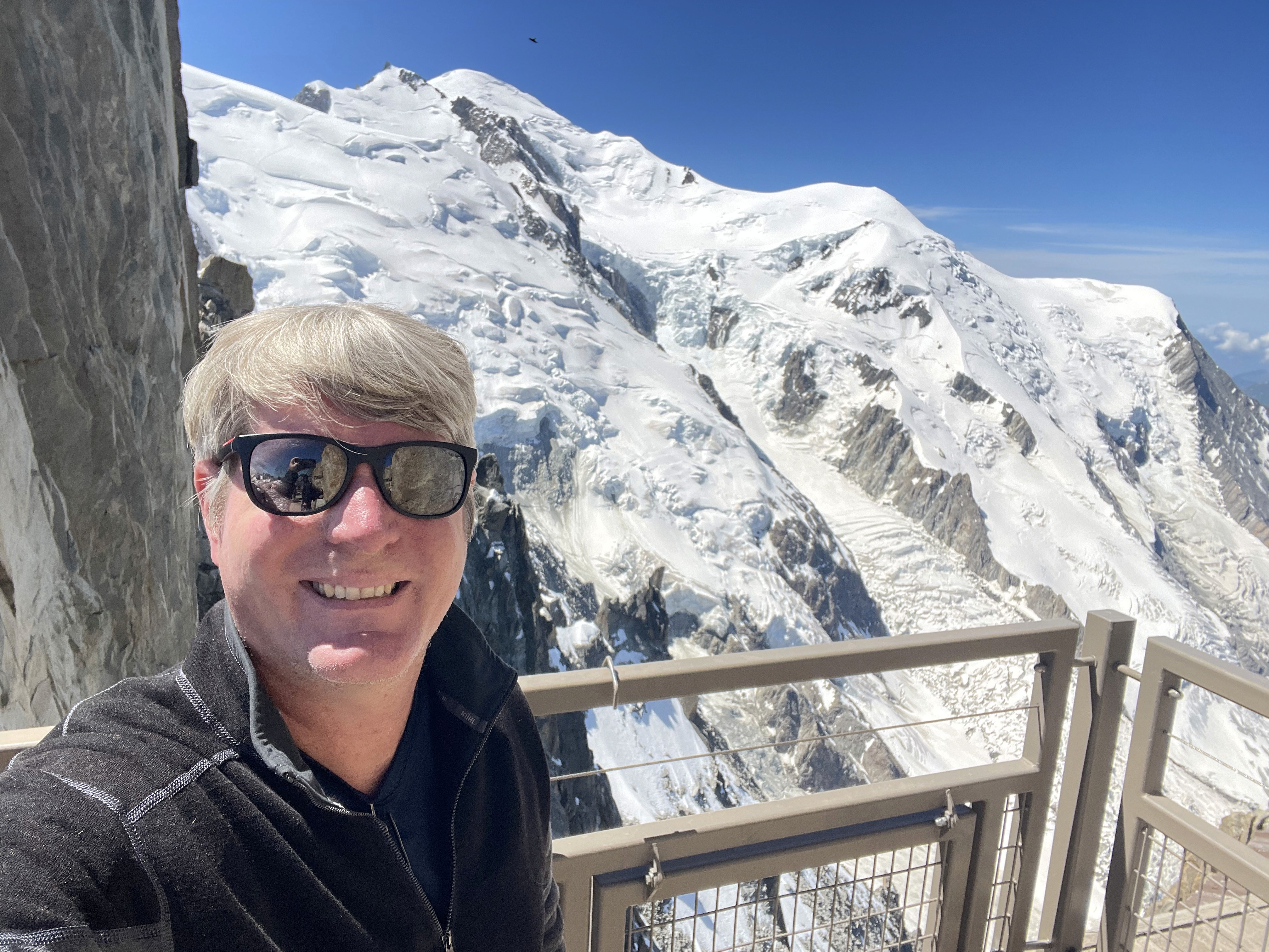 A close selfie of Brian Peddie in sunglasses with snow-covered mountains behind him.