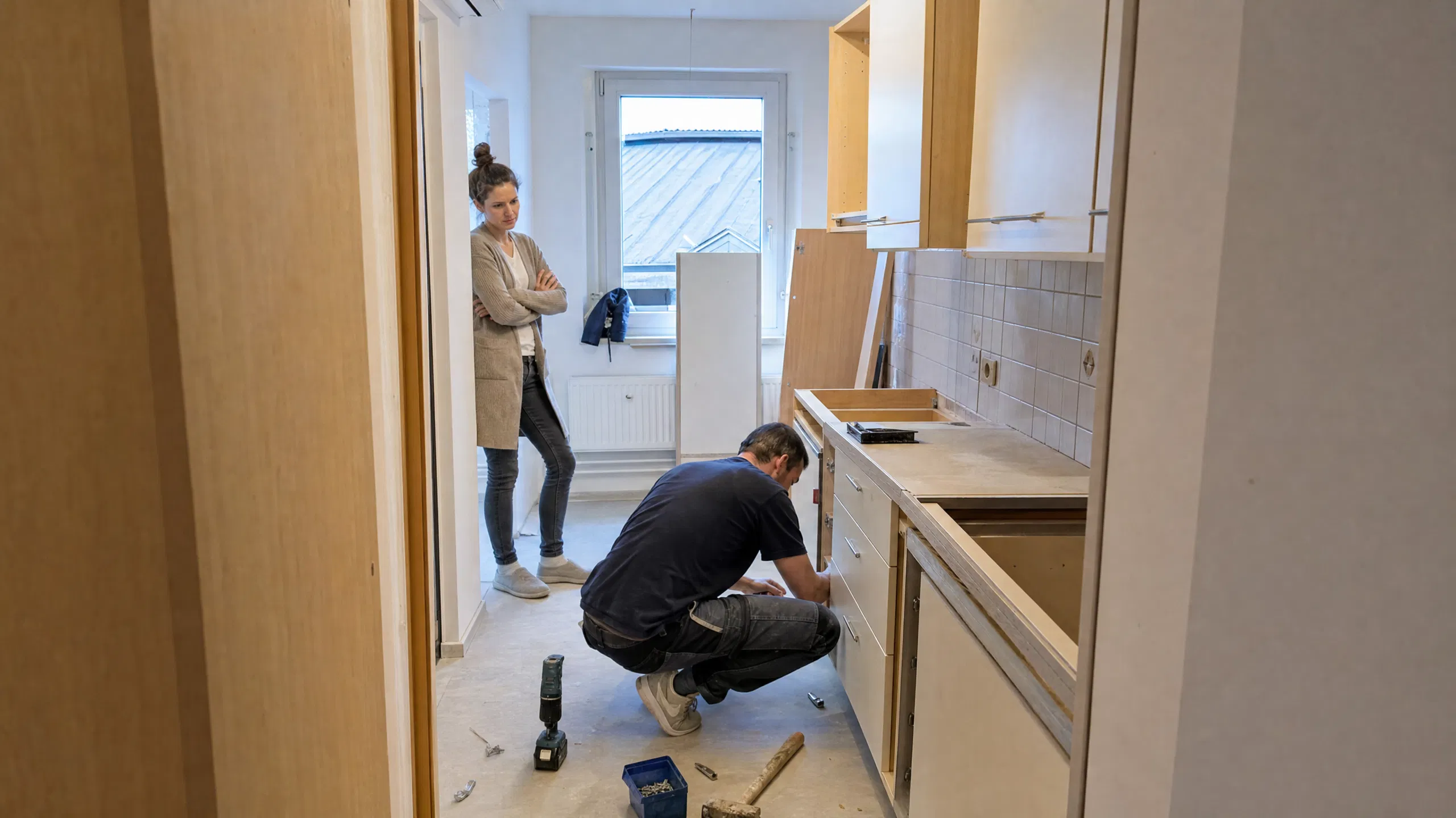 Ein Handwerker baut in einer österreichischen Mietwohnung eine neue Küche ein, während die Mieterin nachdenklich von der Türe aus zuschaut.