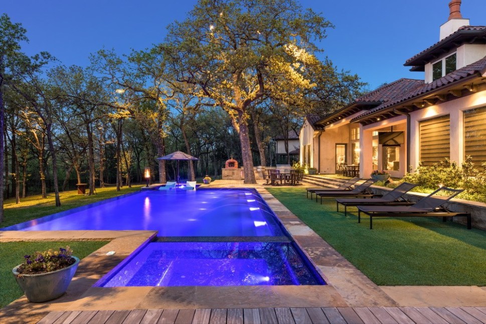 Lakewood estate in Dallas at twilight, featuring a modern pool with blue LEDs, surrounded by professionally installed artificial turf and extensive landscape lighting by Elite Landscaping DFW, including dramatic uplighting on mature trees.