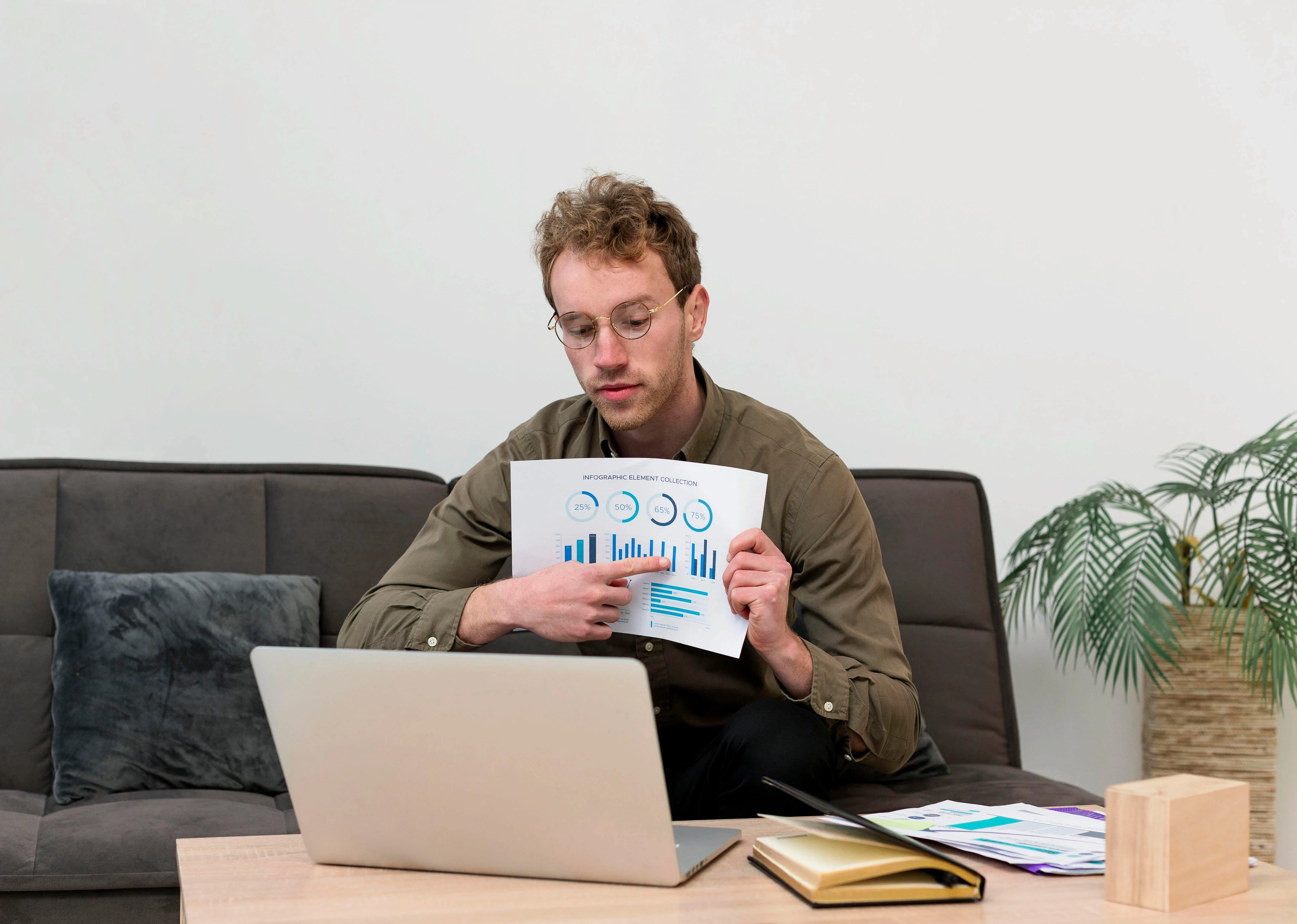 A young man sitting on a sofa and presenting a printed data chart during a video call on his laptop while working from home.