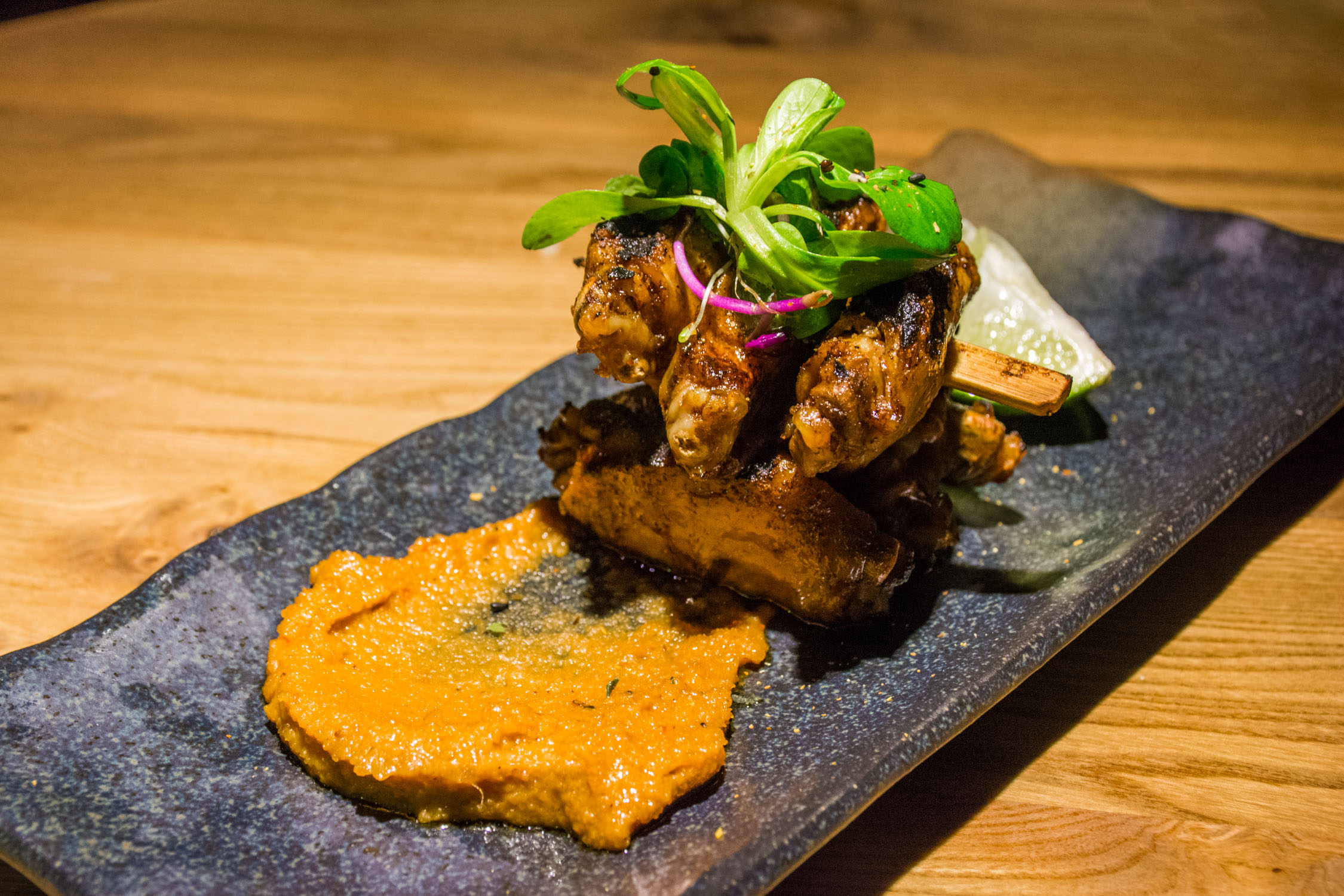 A piece of seared meat topped with greens and red onion sits on a dark stone platter next to an orange puree.