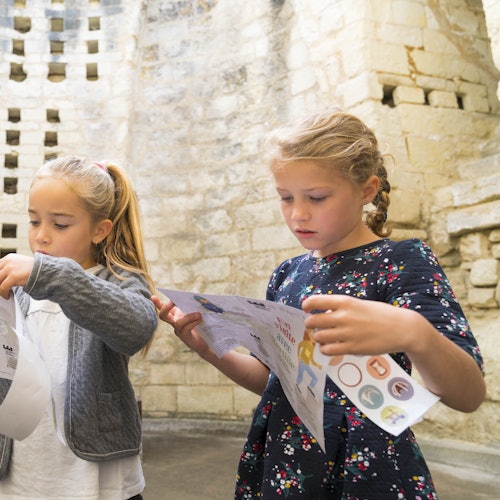 Dos chicas jóvenes examinan papeles con dibujos, de pie frente a un viejo muro de piedra.