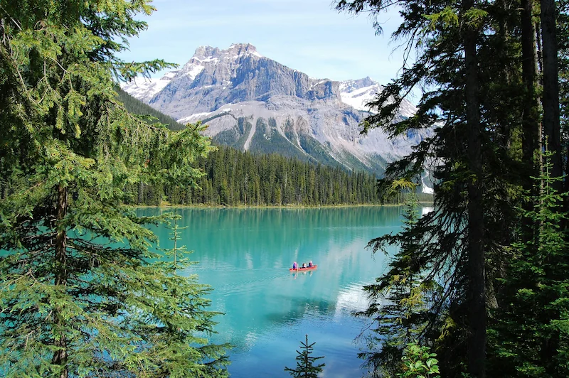Turquoise alpine lake in Canada with evergreen forest, mountain peaks, and a small canoe on calm water.