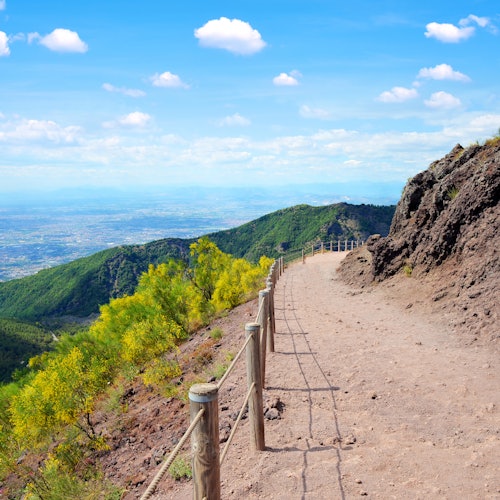 A dirt hiking trail with a wooden fence winds along a mountainside, offering a view of green hills and a blue sky with clouds.