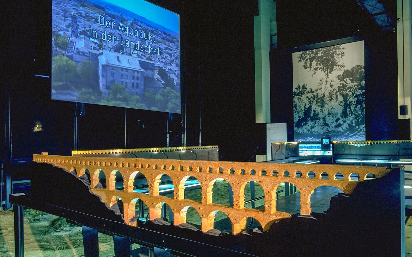 Model of Pont du Gard aqueduct in museum exhibit with cinema screen in background.