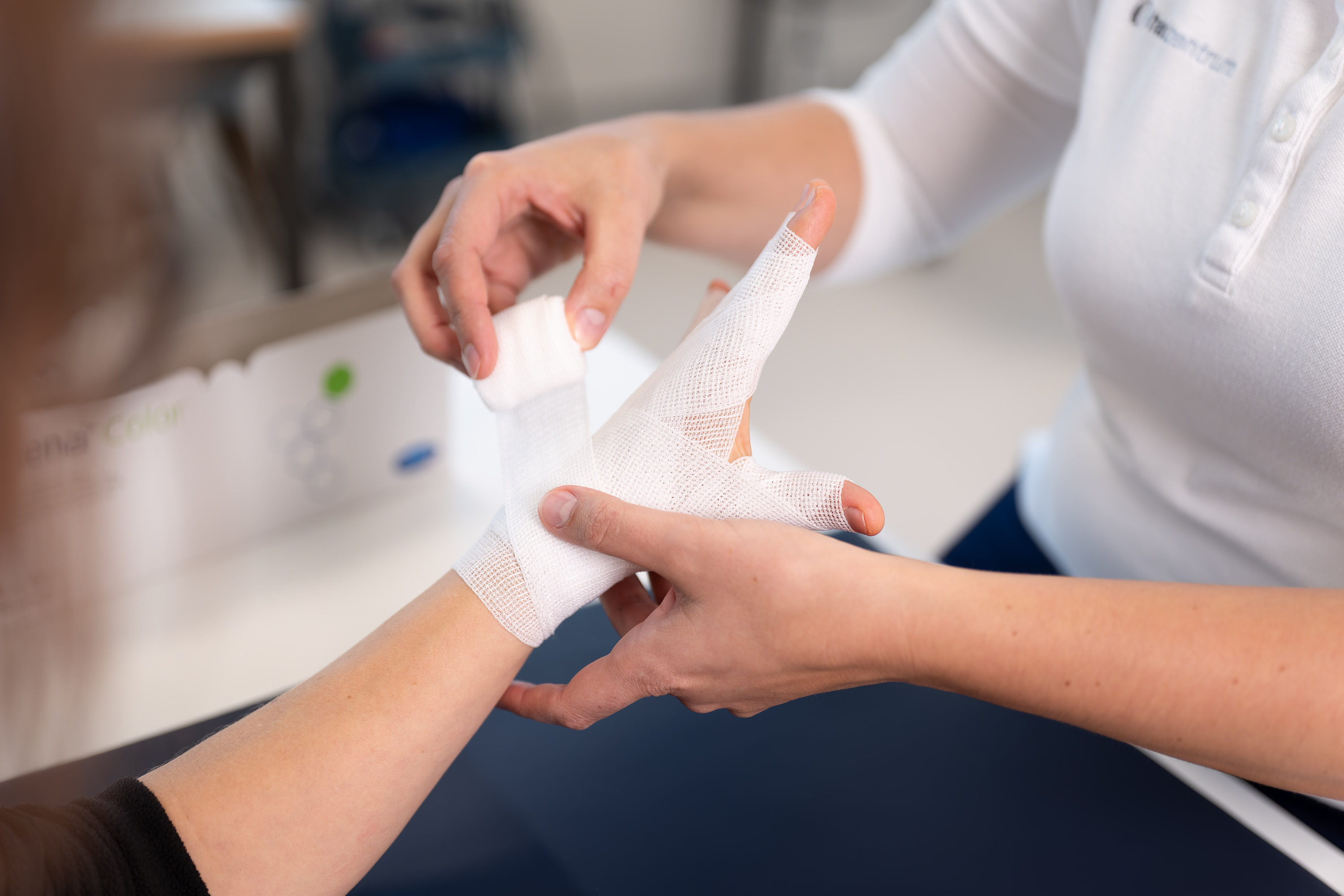 Eine Handtherapeutin umwickelt die Hand und die Finger eines Patienten mit einer weissen Mullbinde und versorgt so eine Verletzung im klinischen Umfeld.