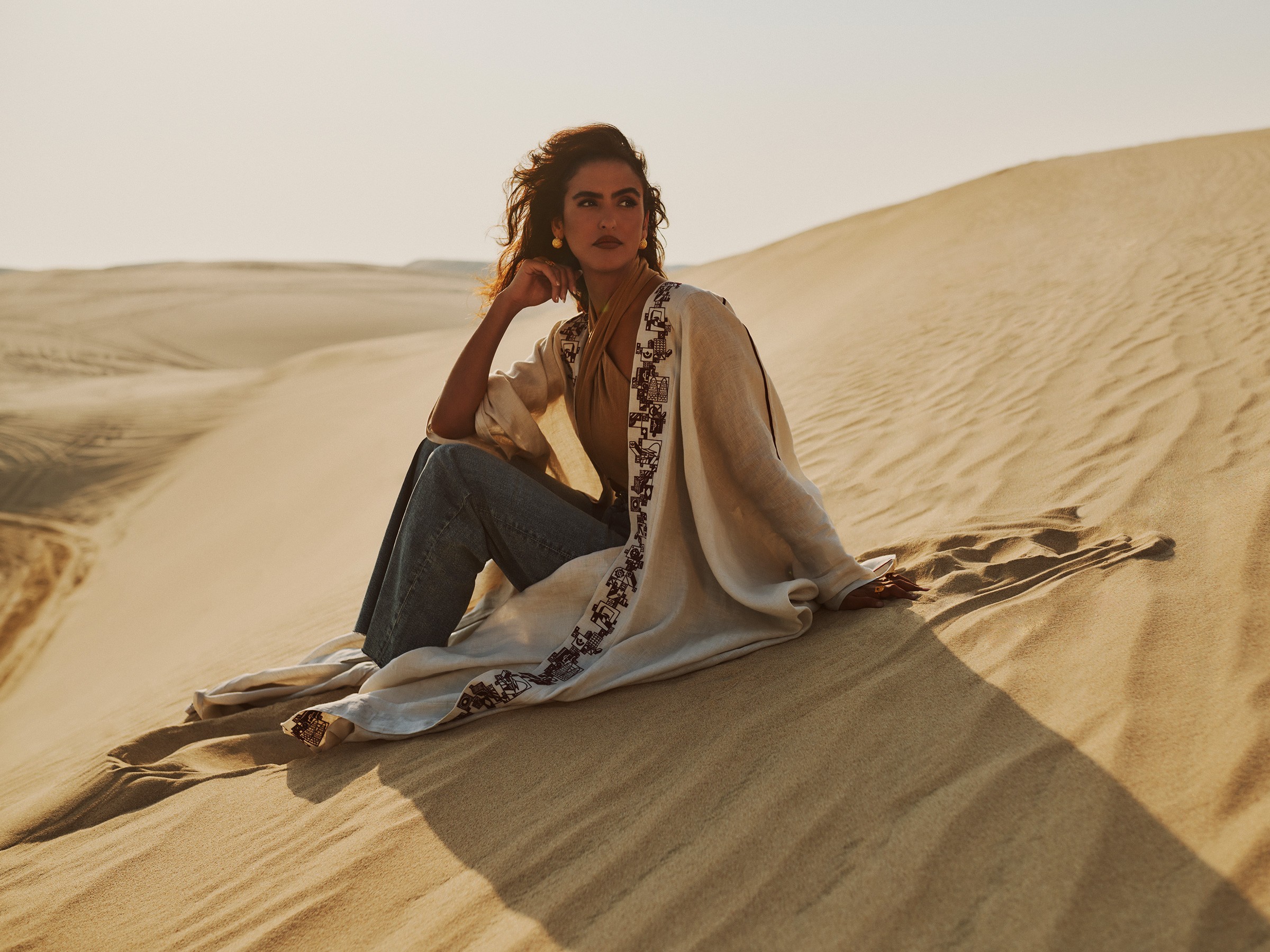 A model seated on a sand dune at golden hour in an ivory abaya with dark embroidered trim.