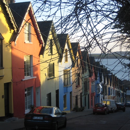 Rangée de maisons colorées sur une rue en pente avec des voitures garées à côté ; de l'eau et un terrain lointain visibles en arrière-plan.