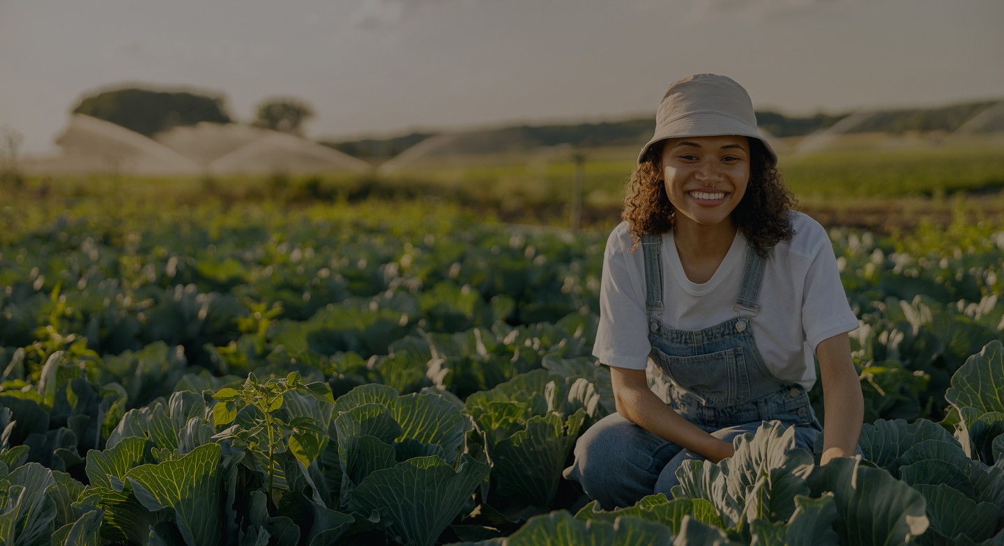 A smiling young woman wearing a bucket hat and denim overalls crouching in a crop field at sunset.