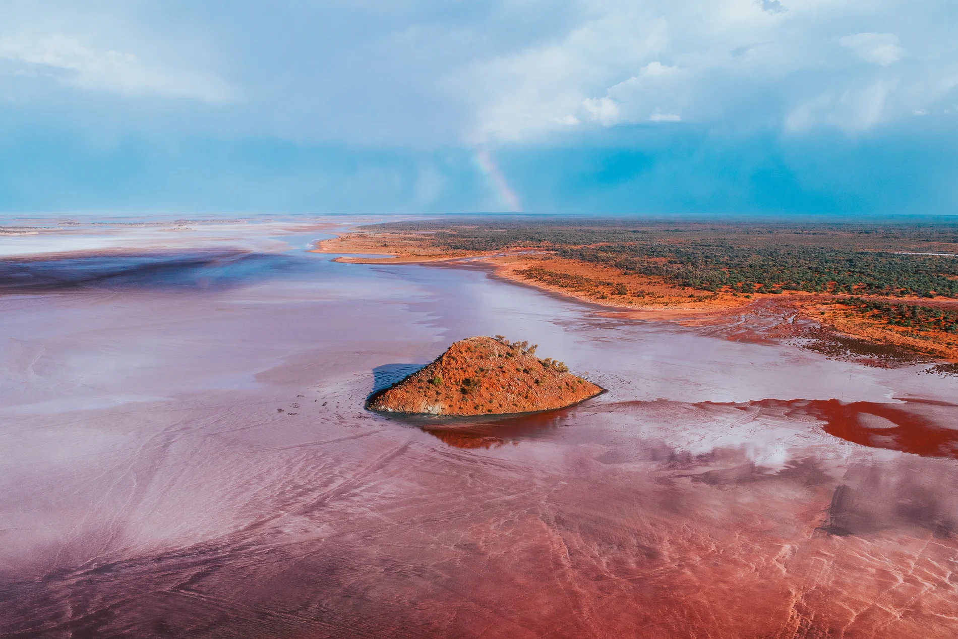 Aerial of a low island in a pink salt lake under a blue sky with rain in the distance