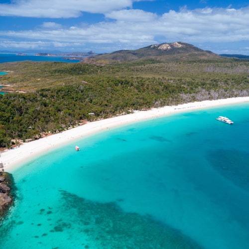 從空中俯瞰潔白的沙灘,碧藍的海水、郁郁蔥蔥的綠色森林山丘,以及在半雲的天空下靠近岸邊的船隻。