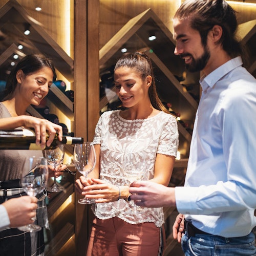 Tři lidé se usmívají, zatímco čtvrtá osoba nalévá víno do jejich sklenic ve vinném sklípku s dřevěnými regály a lahvemi.