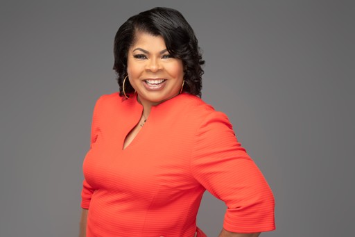 April Ryan with medium-length dark hair smiles confidently, wearing a bright orange dress with three-quarter sleeves. She stands against a plain gray background, posing with one hand on her hip.