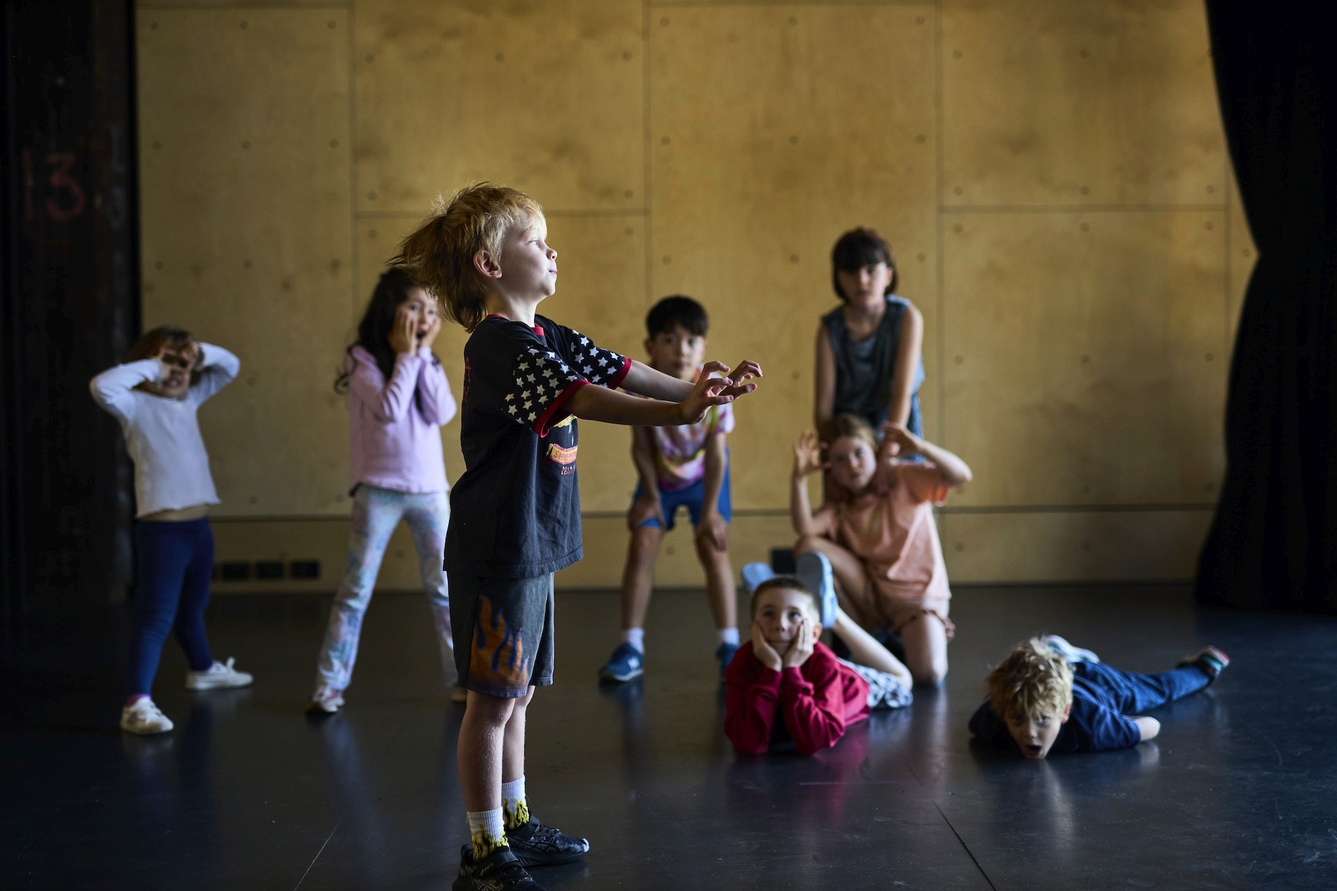 A group of children perform dramatic poses in a studio, with one child standing forward while others crouch, lie down, or gesture behind him.