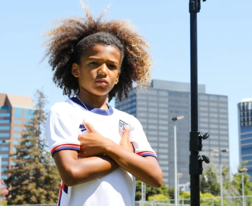 Young soccer player in a USA jersey standing confidently with crossed arms, with a Reeplayer camera set up in the background