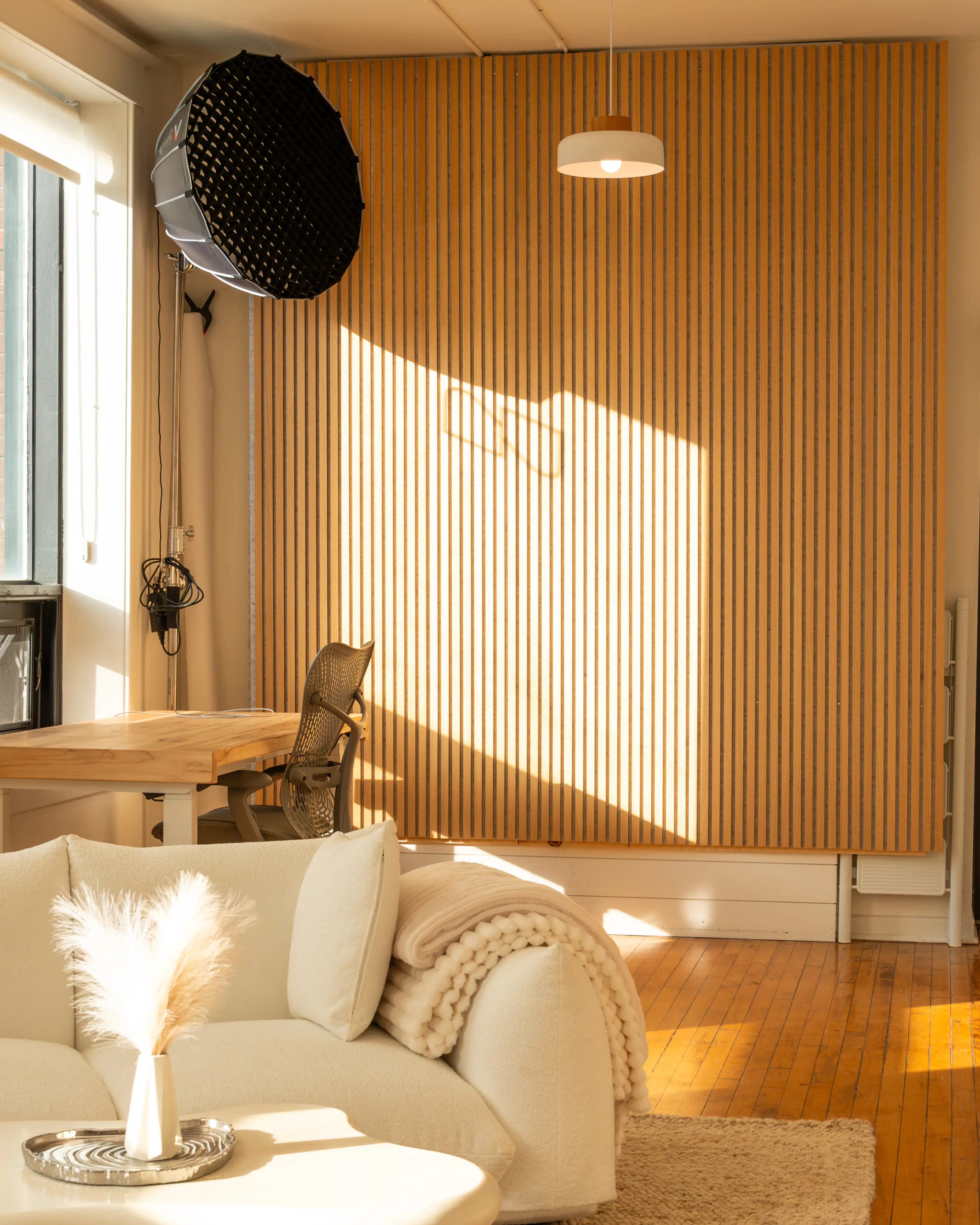 Node Studio’s open-concept layout with warm hardwood floors, natural light pouring through large windows, a cozy white couch on a textured rug, and a slatted wood accent wall creating a bright and inviting atmosphere