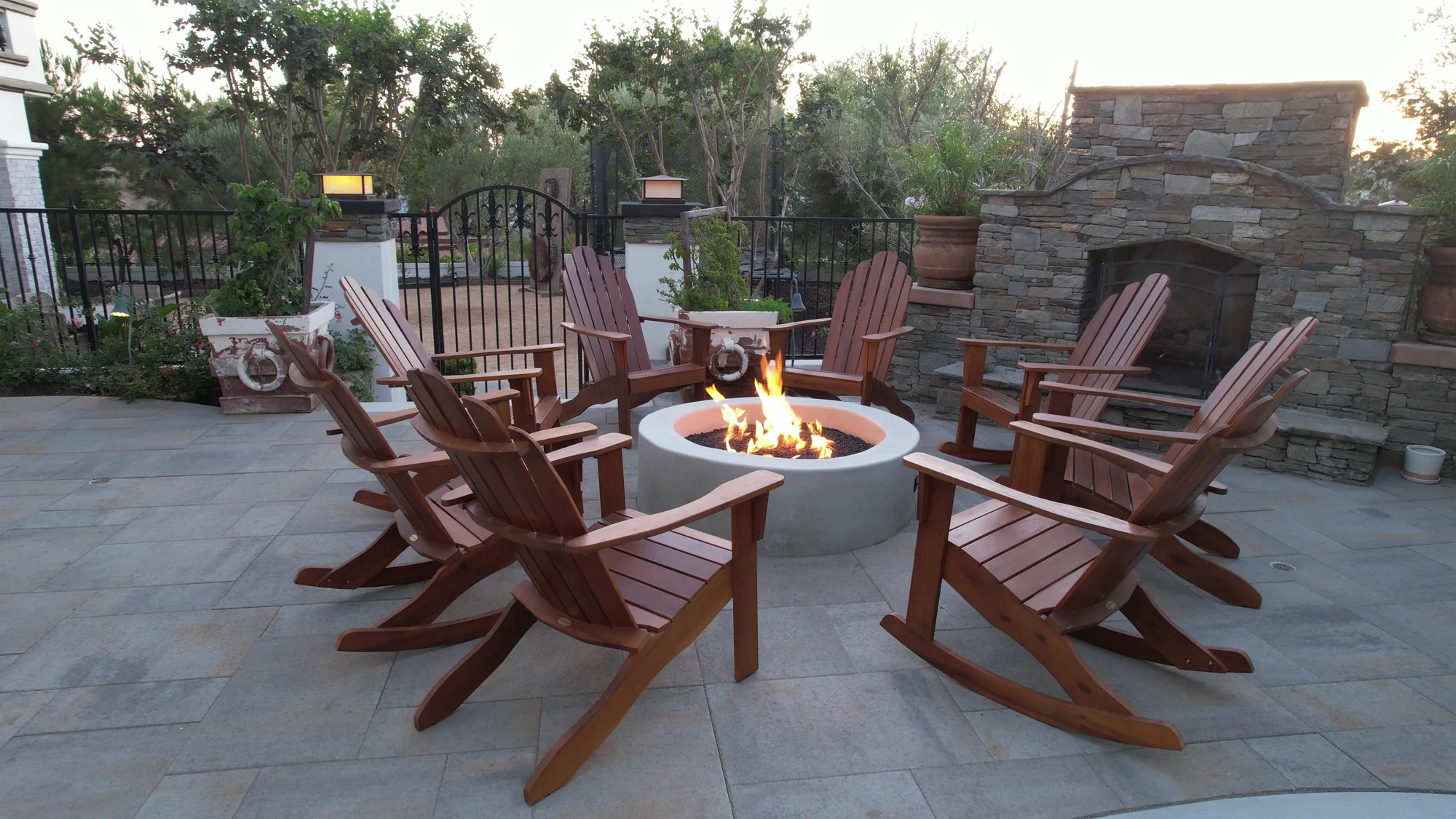 Wooden chairs surround a lit round fire bowl on a paved patio beside a stone fireplace.