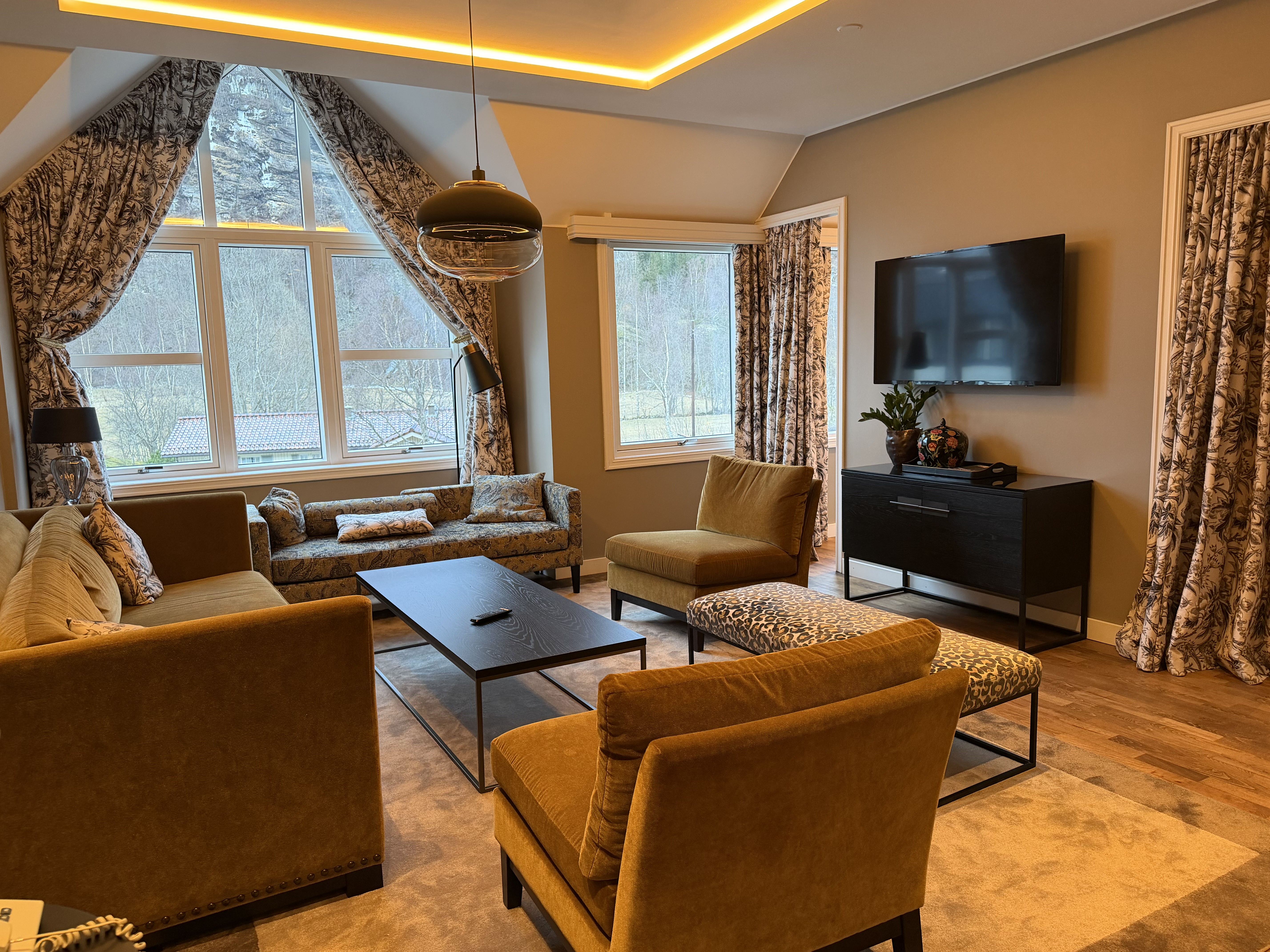A cozy living room with a large window showcasing a lush green landscape, furnished with a green velvet sofa, cream armchair, round coffee table, and decorative pillows, complemented by artwork and a standing lamp.